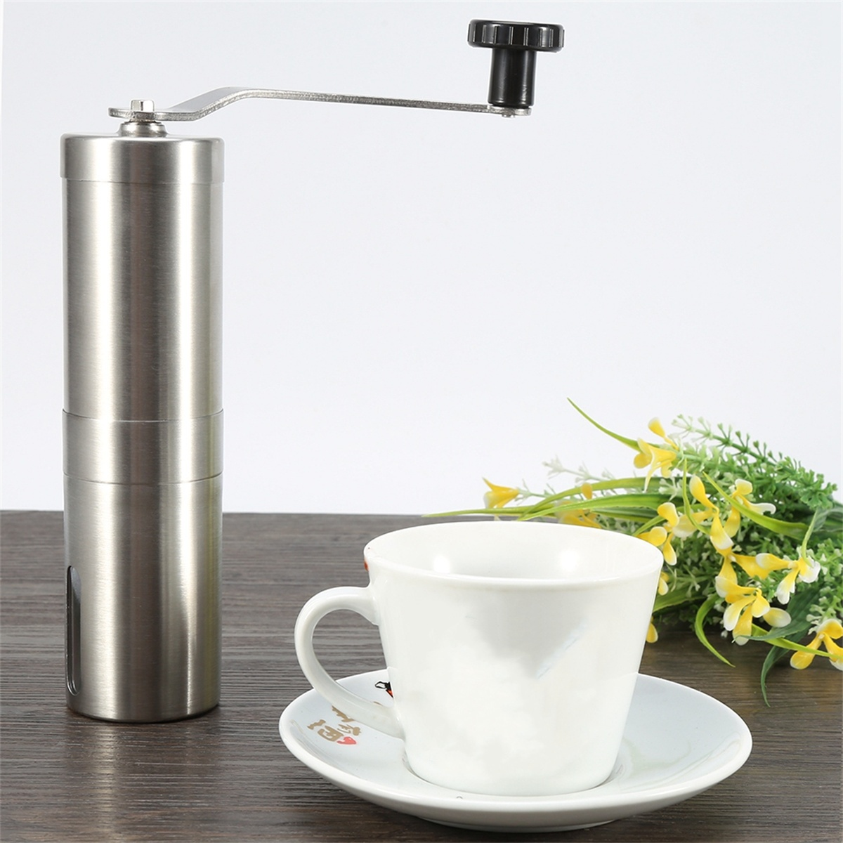 Eine Kaffeemühle aus Edelstahl, eine weiße Tasse und Untertasse sowie einige Blumen. Auf einer Holzoberfläche.