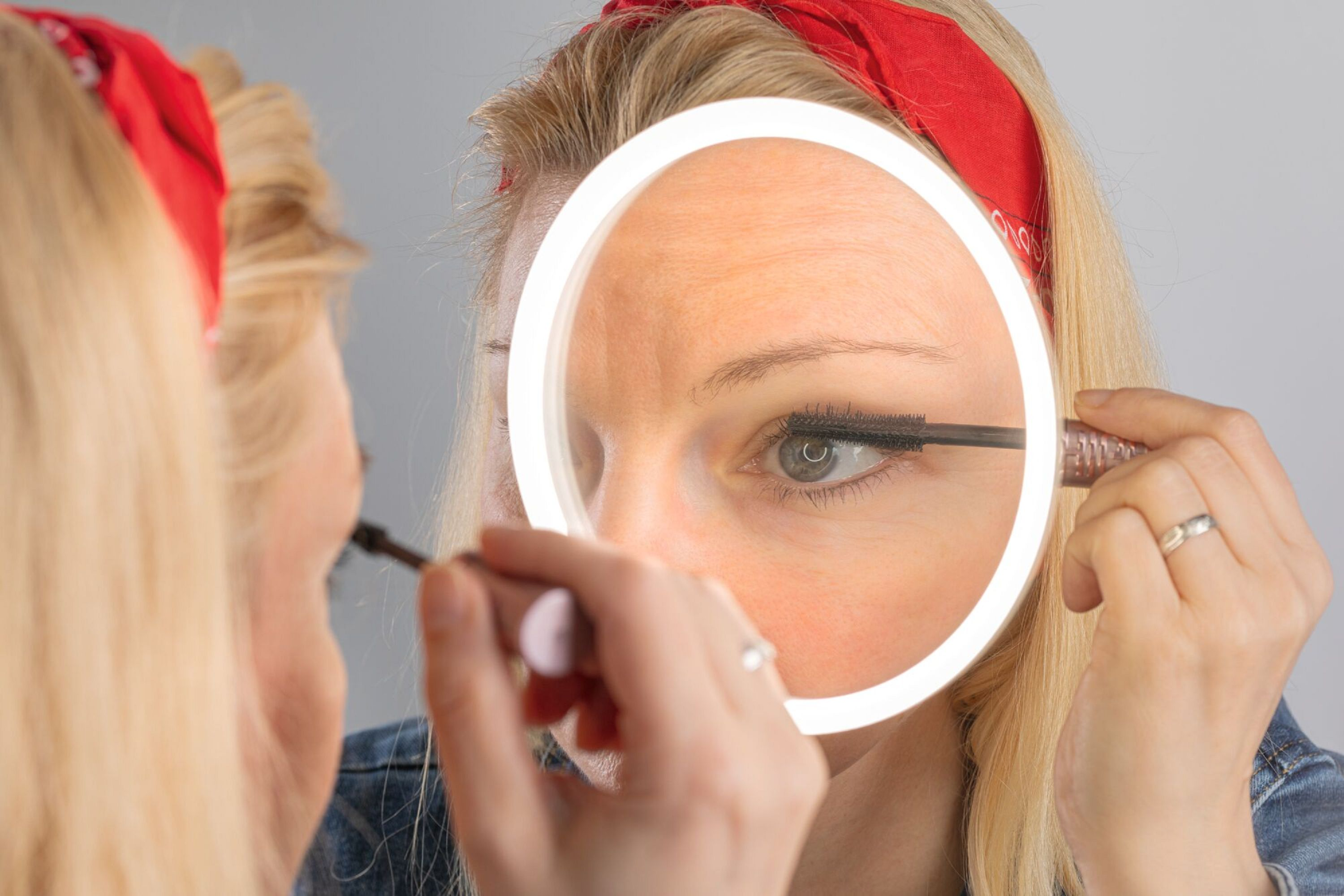 Frau trägt Mascara mit einem Spiegel auf. Sie trägt ein rotes Stirnband. Hintergrund ist eine graue Wand.
