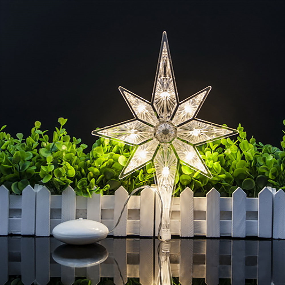Ein beleuchteter Stern-Baumschmuck sitzt über einem weißen Zaun und einer grünen Hecke vor einem schwarzen Hintergrund. Vorne ist ein weißes Gerät.