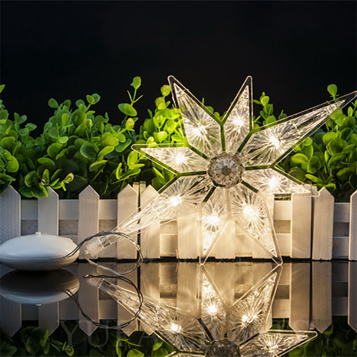 Sternförmiger Weihnachtsbaumschmuck mit Lichtern, spiegelt sich auf der Oberfläche, vor grünen Pflanzen.