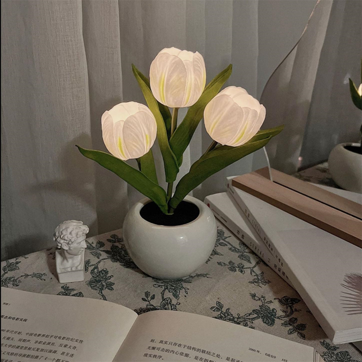 Drei beleuchtete tulpenförmige Lichter in einer weißen Vase auf einem Tisch mit Büchern und einer Statue.