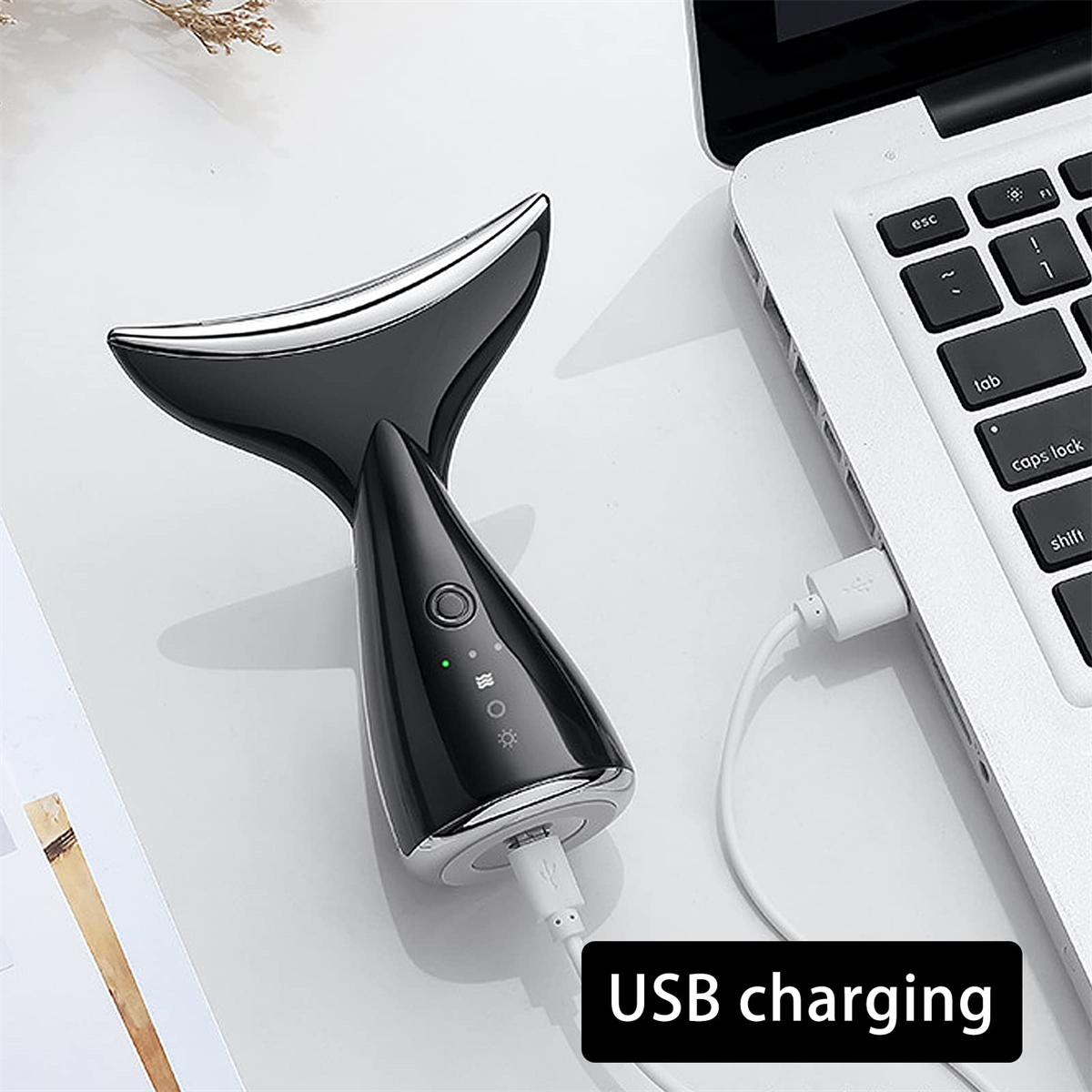 Schwarzes Gesichtsmassagegerät, das über ein USB-Kabel auf einer weißen Oberfläche geladen wird.