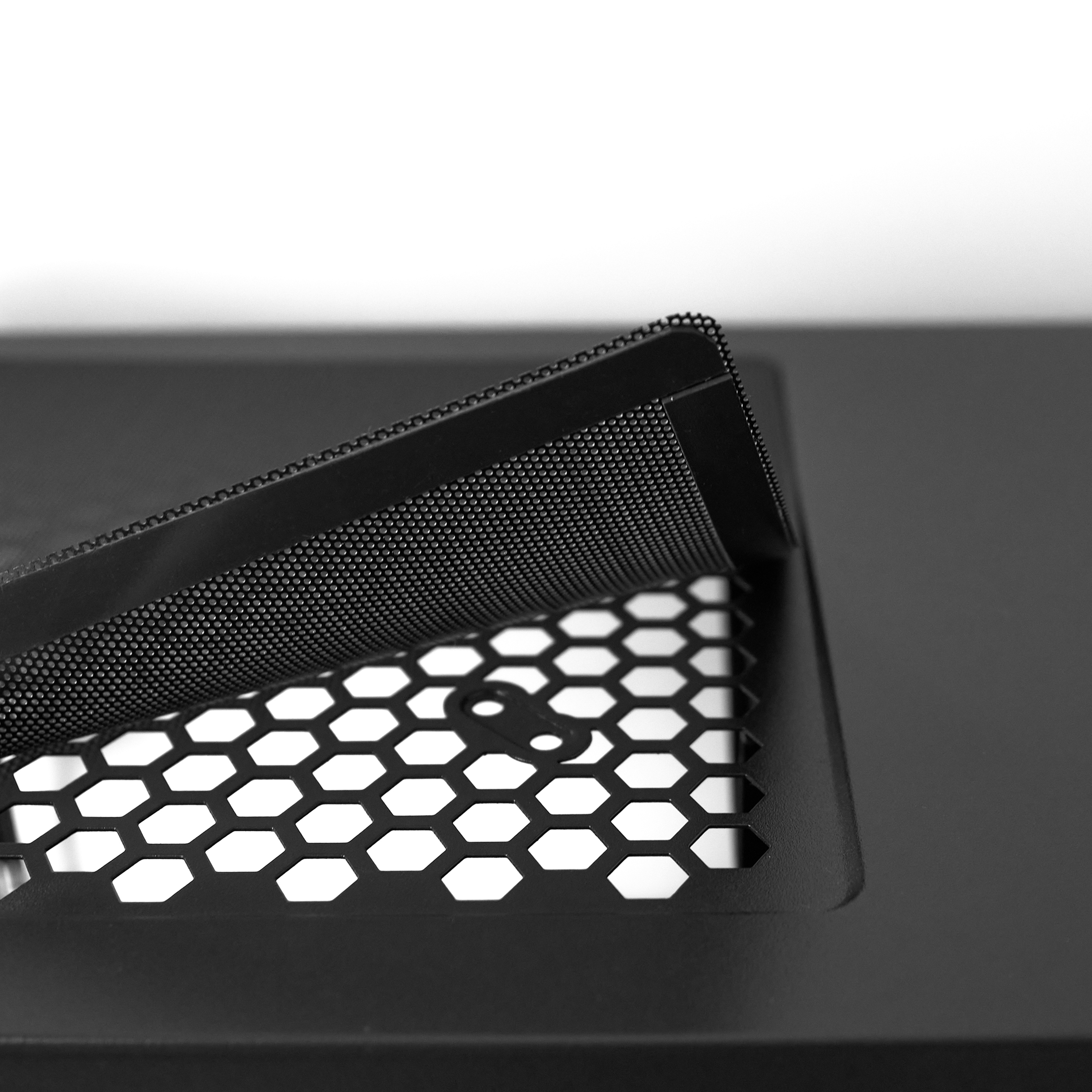 Zbliżenie czarnej metalowej siatki na powierzchni o wzorze plastra miodu. Siatka jest częściowo podniesiona.