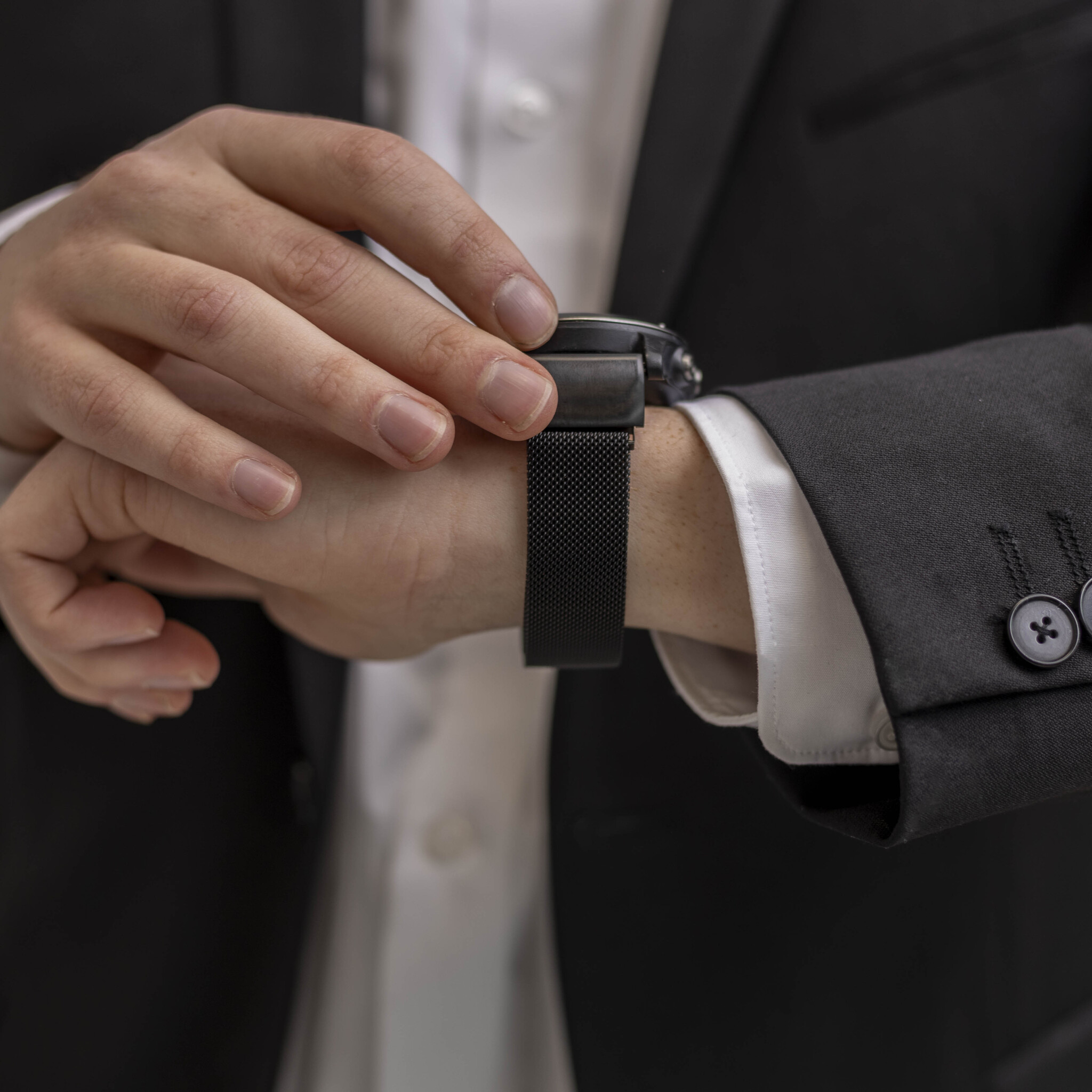Mann stellt eine Uhr mit schwarzem Mesh-Band ein. Er trägt einen schwarzen Anzug und ein weißes Hemd. Fokus auf die Uhr.