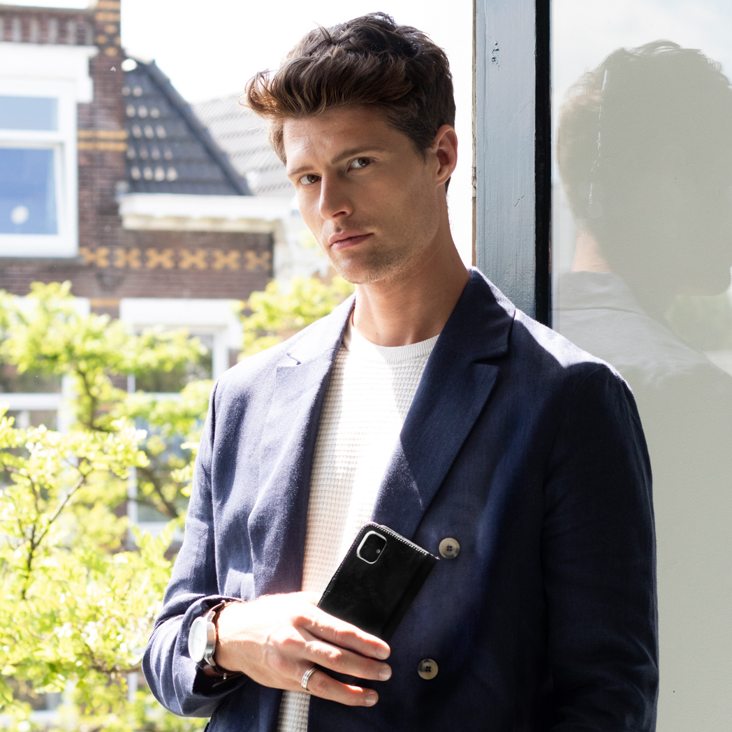 Een man in een blauwe blazer en wit overhemd staat bij een raam en houdt een zwarte telefoonhoes vast.