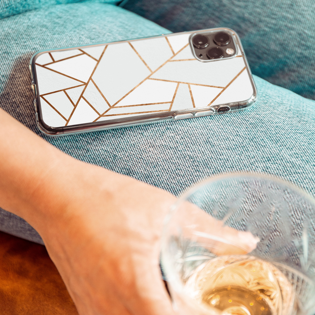 Een telefoon met een witgouden hoesje ligt op een spijkerbroek. Een hand houdt een wijnglas met amberkleurige vloeistof vast.