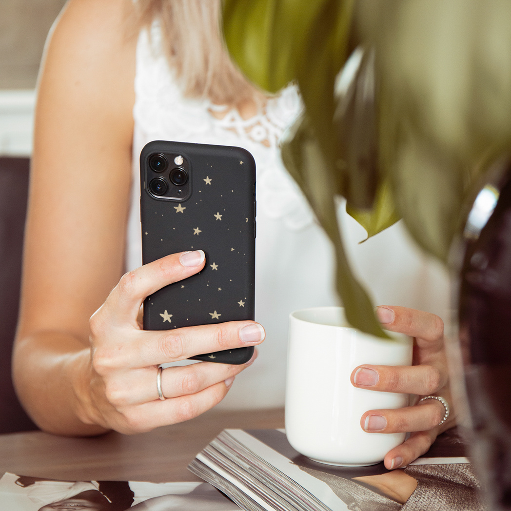 Een vrouw houdt een zwarte telefoon vast met gouden sterren. De telefoon heeft een witte hoes. Ze houdt een witte beker vast.