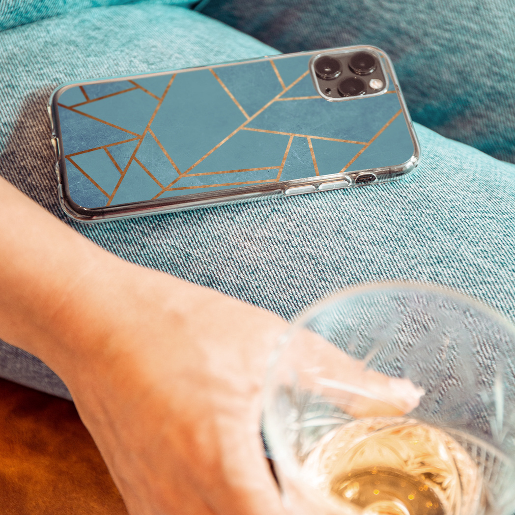 Personne tenant un téléphone avec un design bleu et or, un jean et un verre avec une boisson sur une surface bleue.