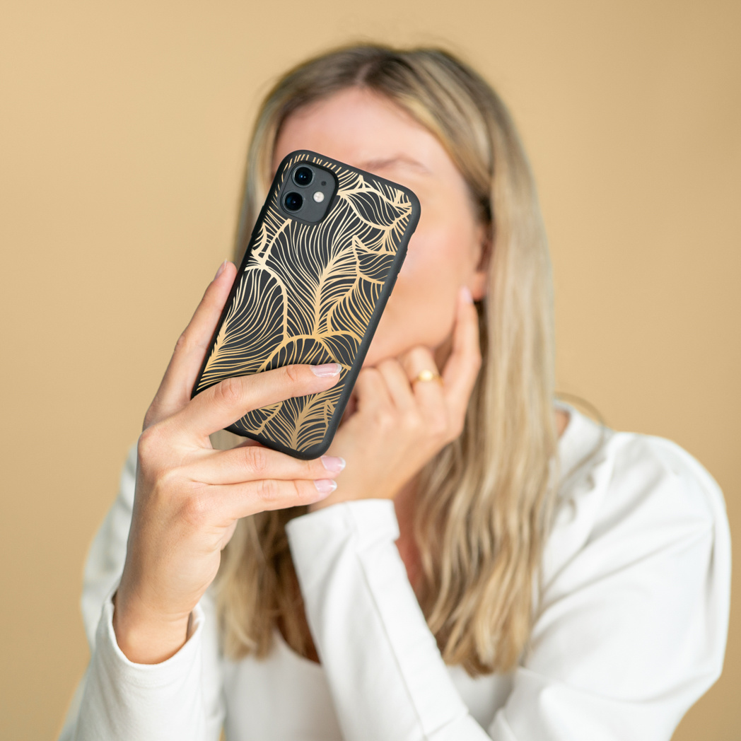 Een vrouw die een zwarte telefoon vasthoudt met een gouden bladerenpatroonhoesje, dat haar gezicht bedekt.