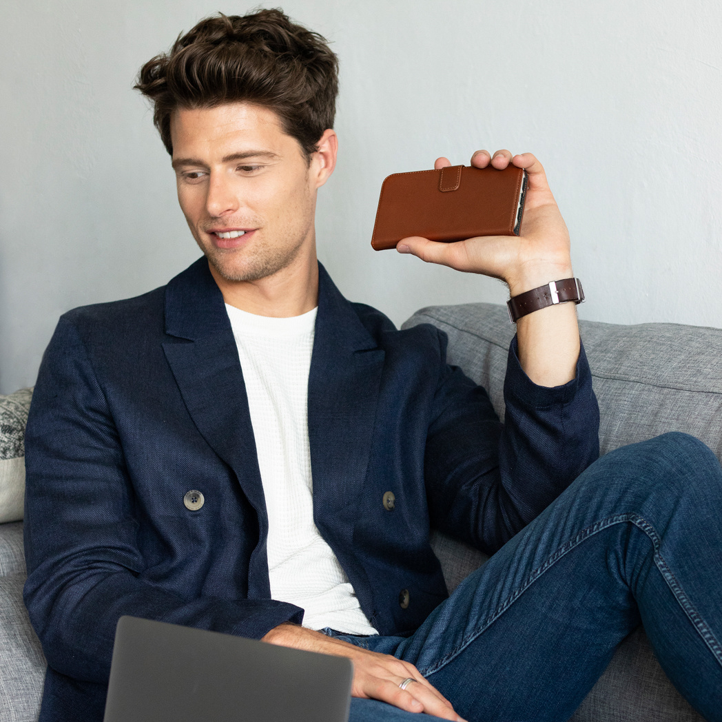 Mann in Blazer hält braune Handyhülle. Er sitzt auf grauem Sofa mit Laptop. Hintergrund ist weiße Wand.