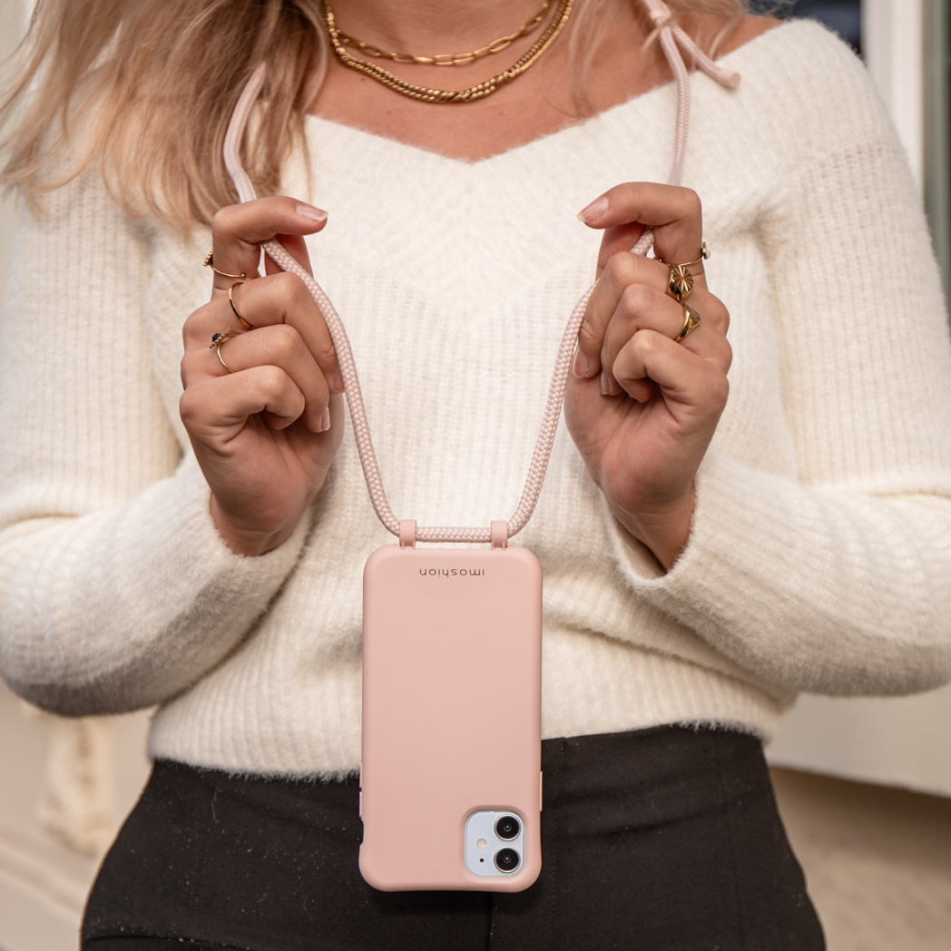 Femme tenant une coque de téléphone rose avec une sangle de cou rose, portant un pull blanc et des bijoux.