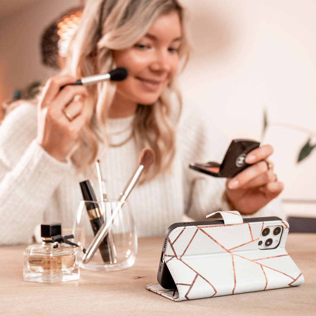Une femme se maquille avec des pinceaux et un téléphone dans un étui blanc et doré sur la table.