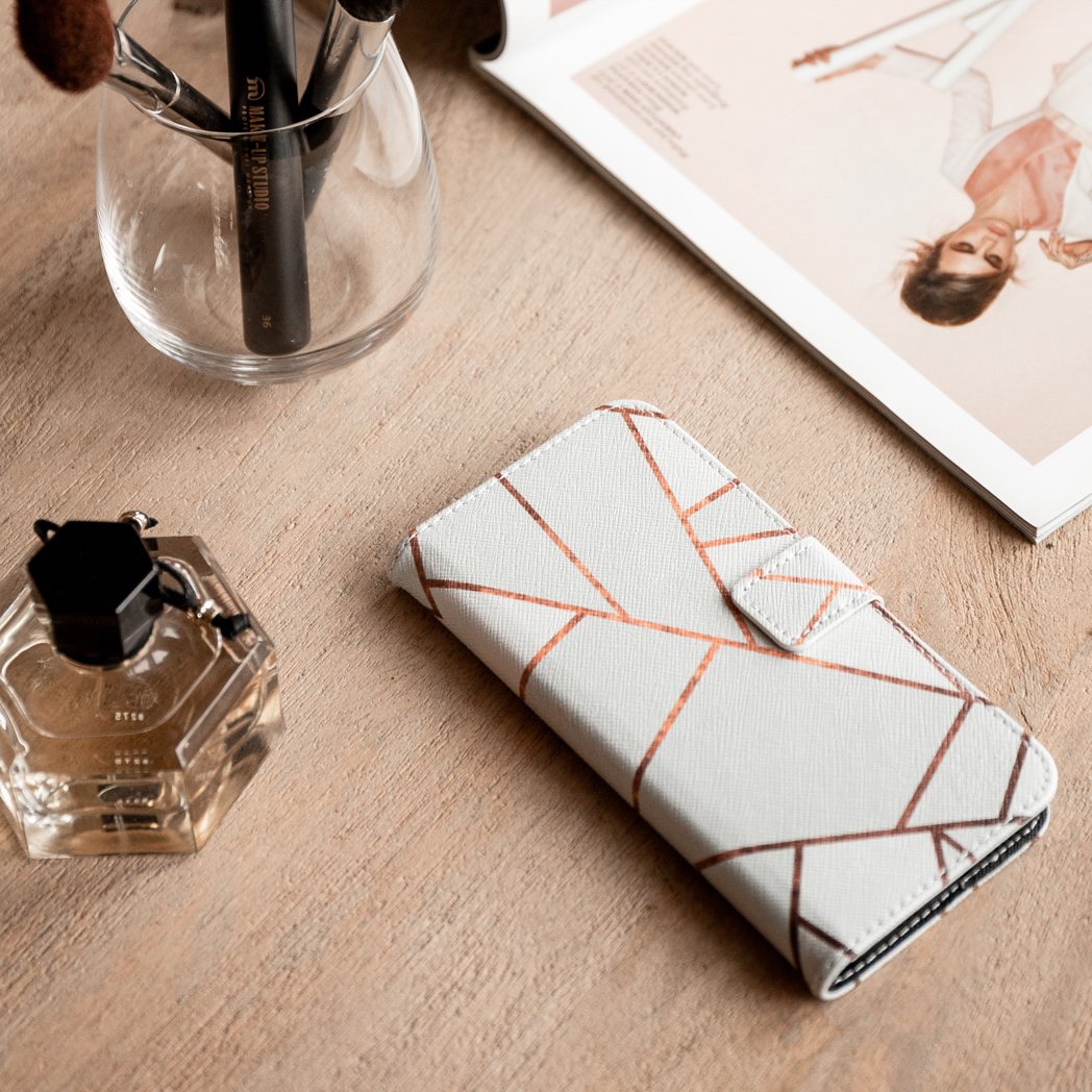 Une coque de téléphone blanche et dorée, des pinceaux de maquillage, du parfum et un magazine sur une table en bois.
