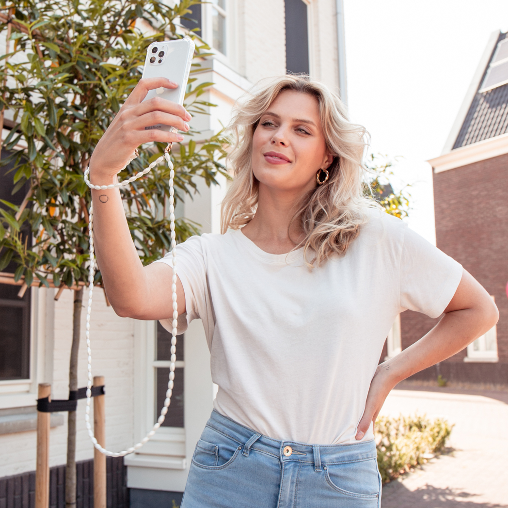 Une femme tenant un téléphone blanc avec une lanière de perles et prenant un selfie, à l'extérieur.