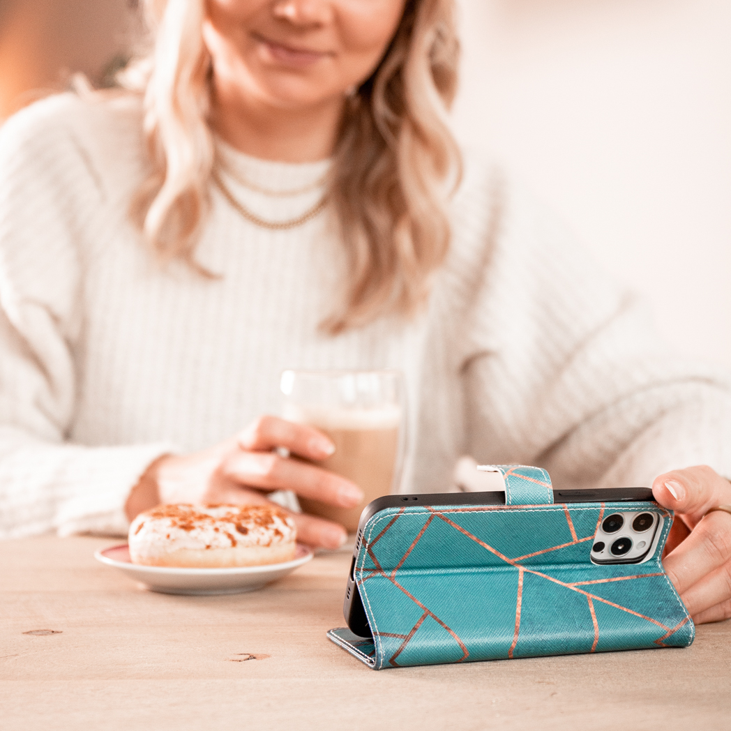 Une femme avec un café et un beignet tient son téléphone dans un étui turquoise et doré.
