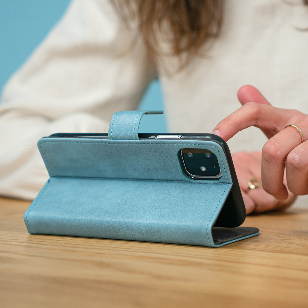 De vinger van een persoon raakt een blauwe telefoonhoes op een houten tafel aan. De hoes is open en ondersteunt de telefoon.