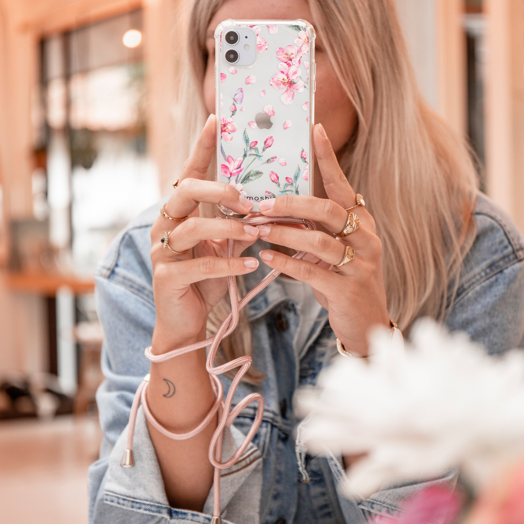 Een vrouw houdt een telefoon met een bloemenhoesje vast. De telefoon heeft een roze koord. Haar handen hebben ringen.