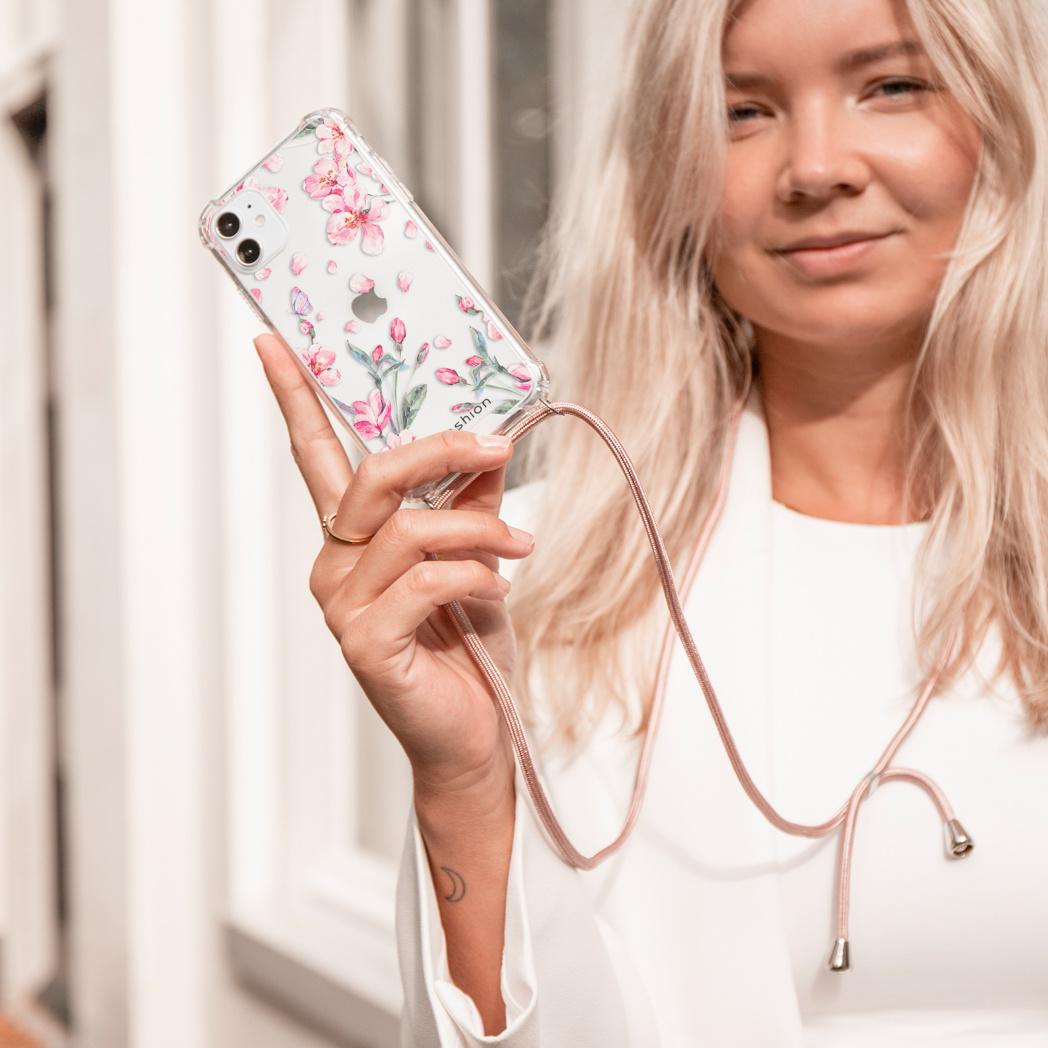 Een vrouw houdt een doorzichtige telefoonhoes met roze bloemenpatroon vast. Met roze koord.