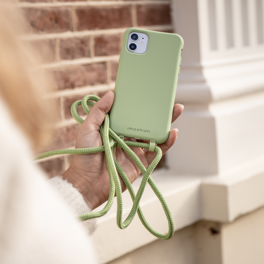 Persoon houdt groene telefoonhoes met groen koord vast, bakstenen muur op de achtergrond.