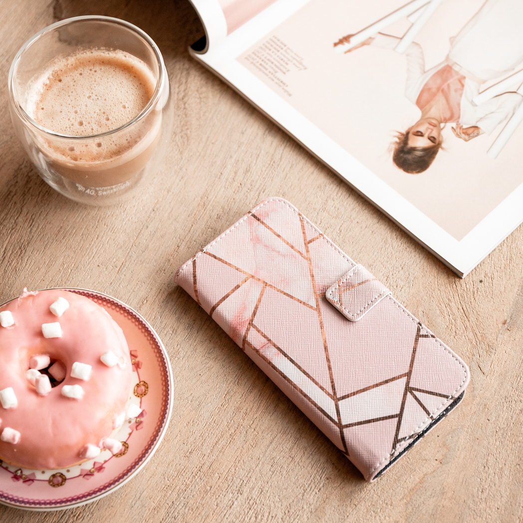 Un téléphone dans un étui rose avec un design géométrique est à côté d'un beignet et d'un café sur une table en bois.