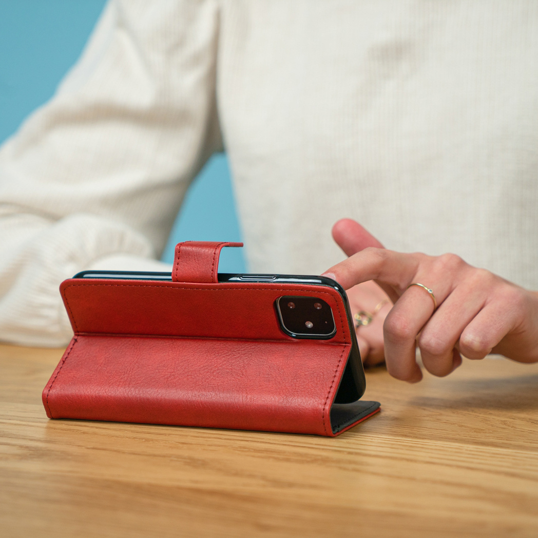 De hand van een persoon raakt een rode telefoonhoes op een houten tafel aan. De telefoon is zwart.