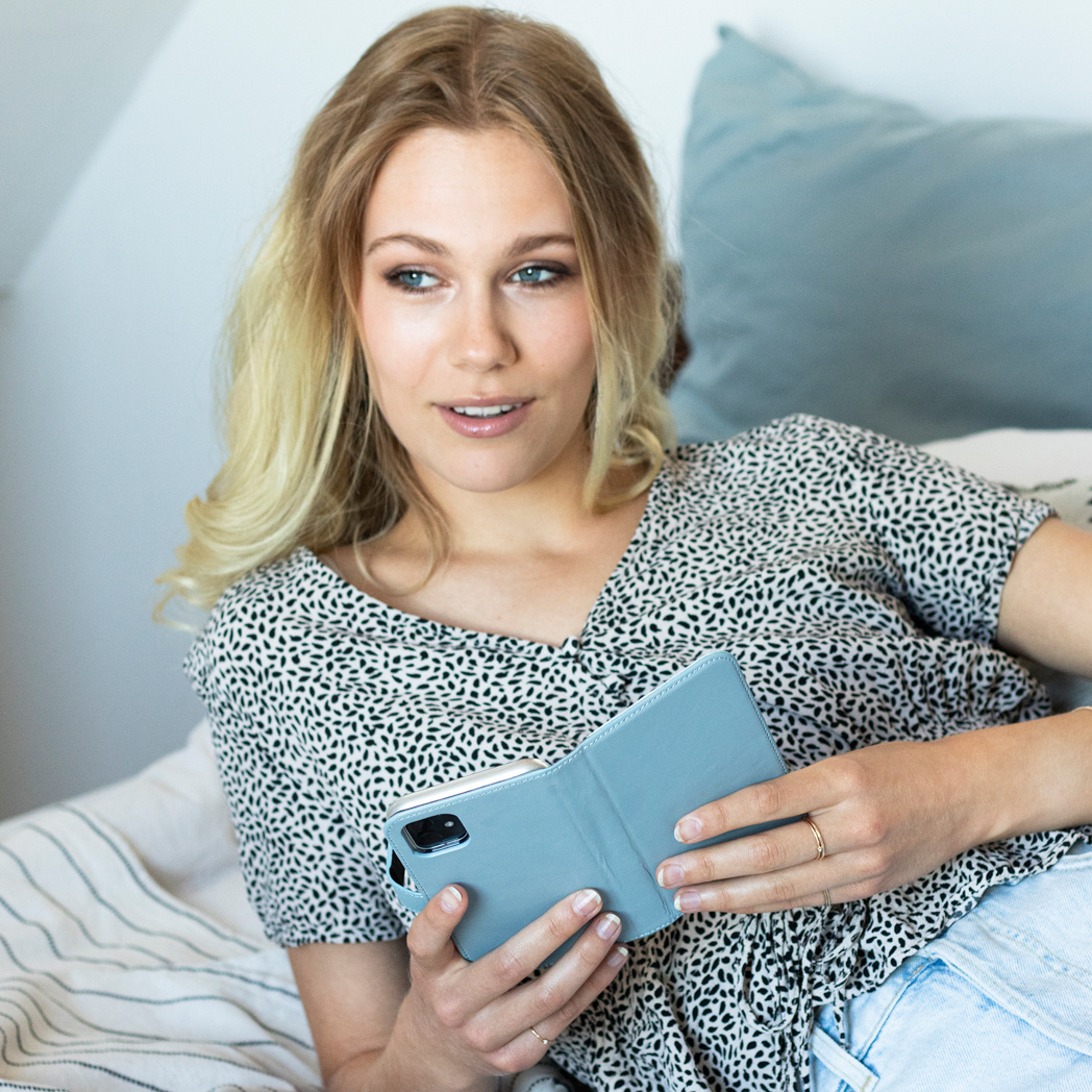 Femme blonde tenant un étui de téléphone, regardant sur le côté. Oreiller et mur en arrière-plan.