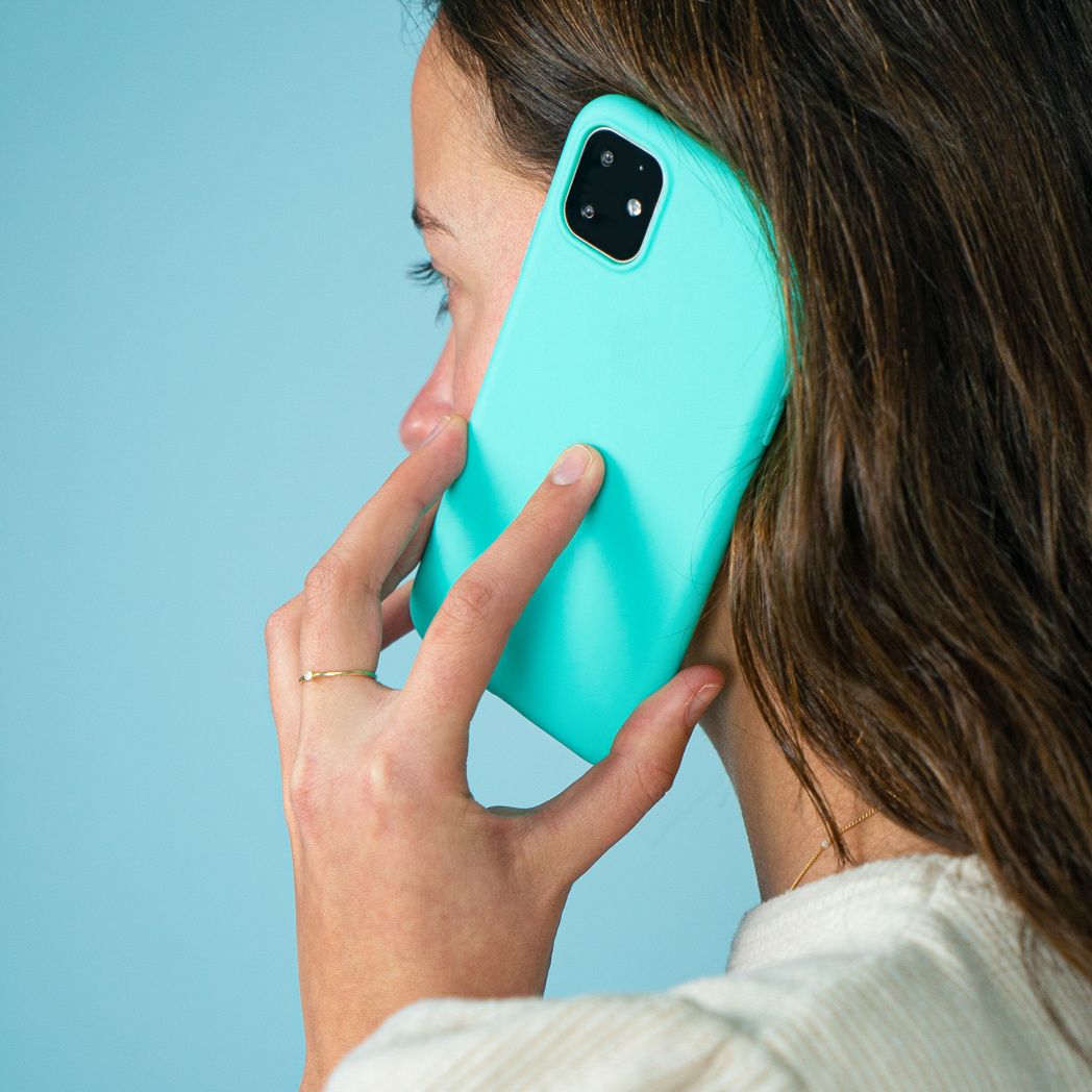 Vrouw houdt een turquoise telefoon tegen haar oor, close-up. Ze draagt een witte trui en een gouden ring. Blauwe achtergrond.