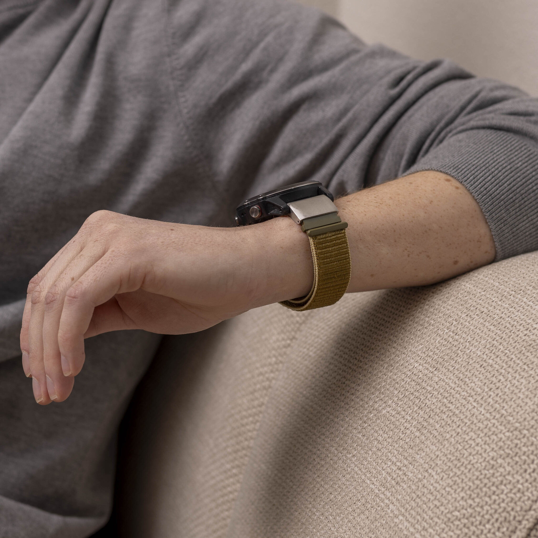 Das Handgelenk einer Person mit einer Uhr und einem grünen Uhrenarmband auf einem beigen Sofa.