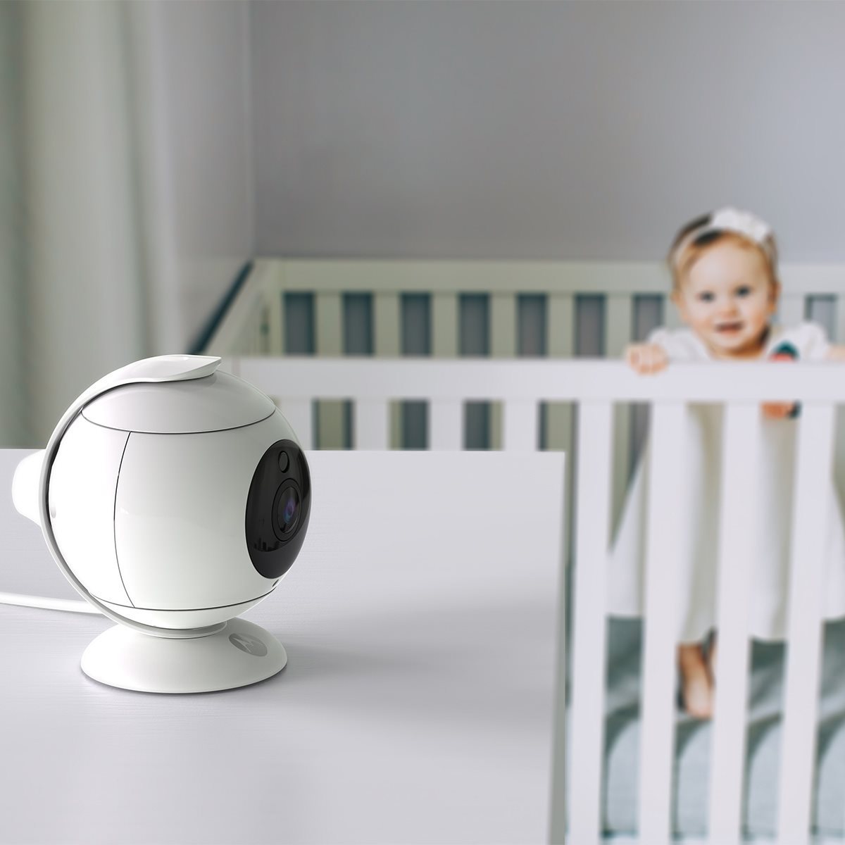 A white baby monitor camera on a table, facing a crib where a baby is smiling.