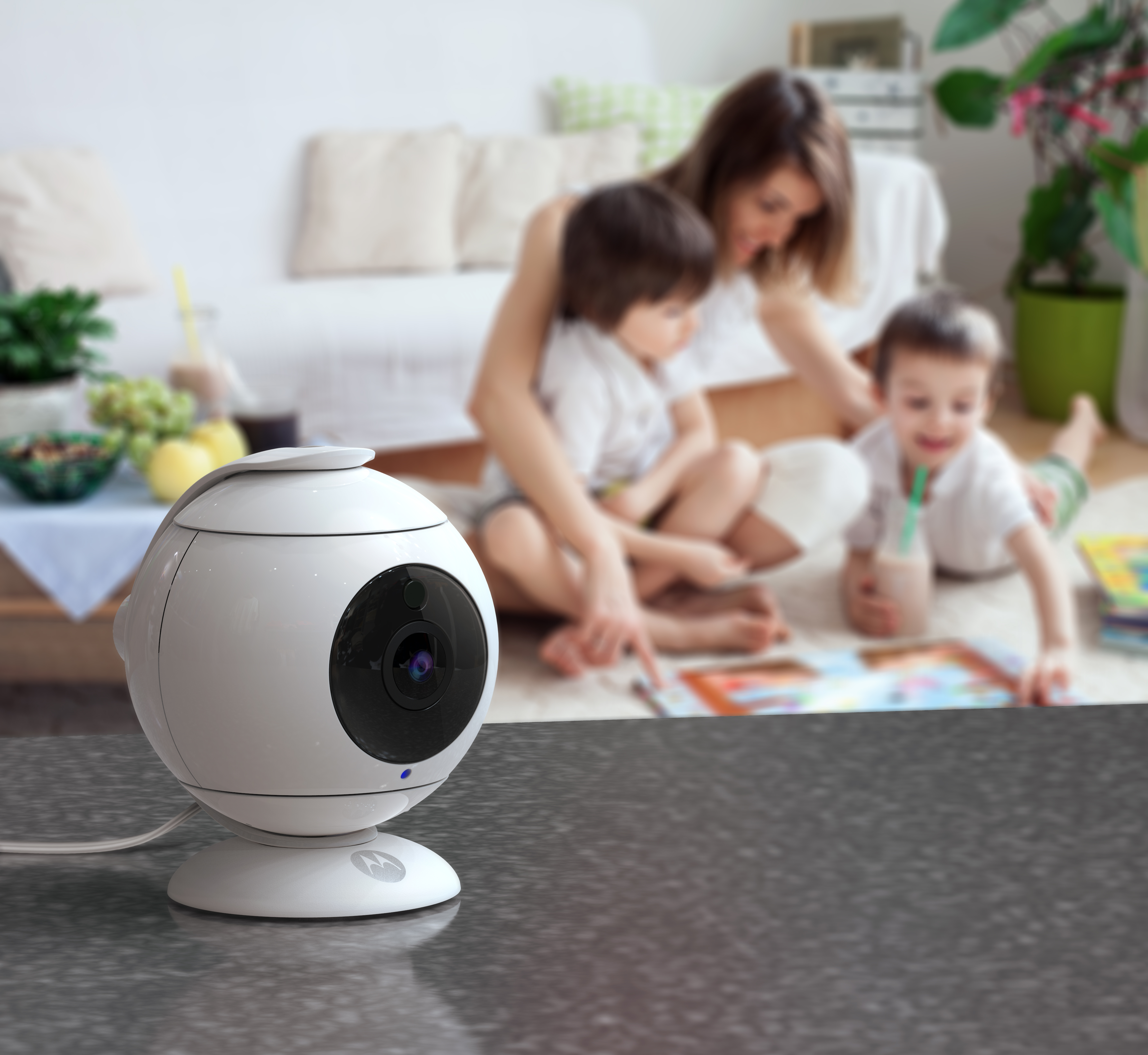 White baby monitor on a dark surface. A family is blurred in the background playing.