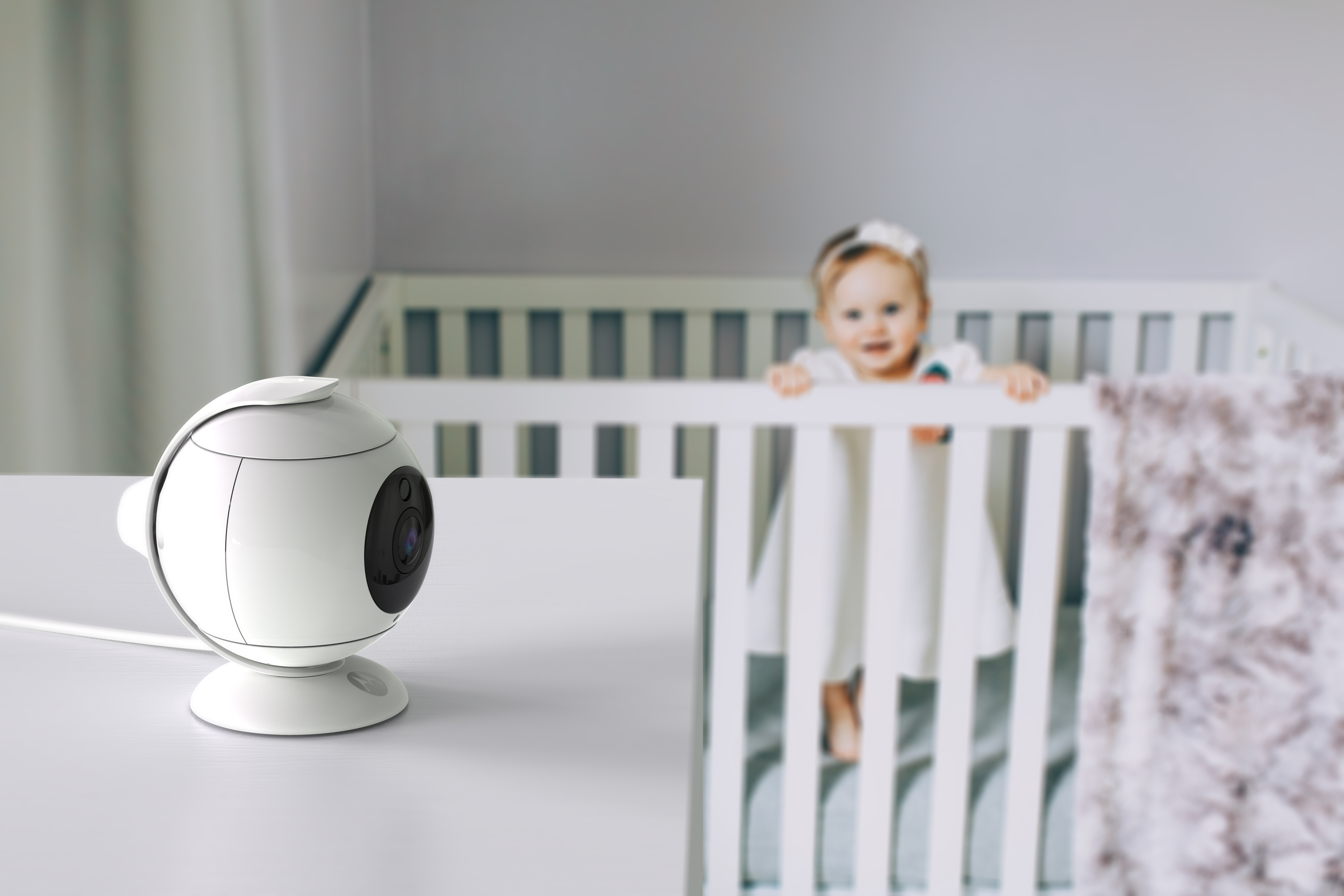 Baby monitor on a white table with a baby in a crib in the background. The baby is smiling.