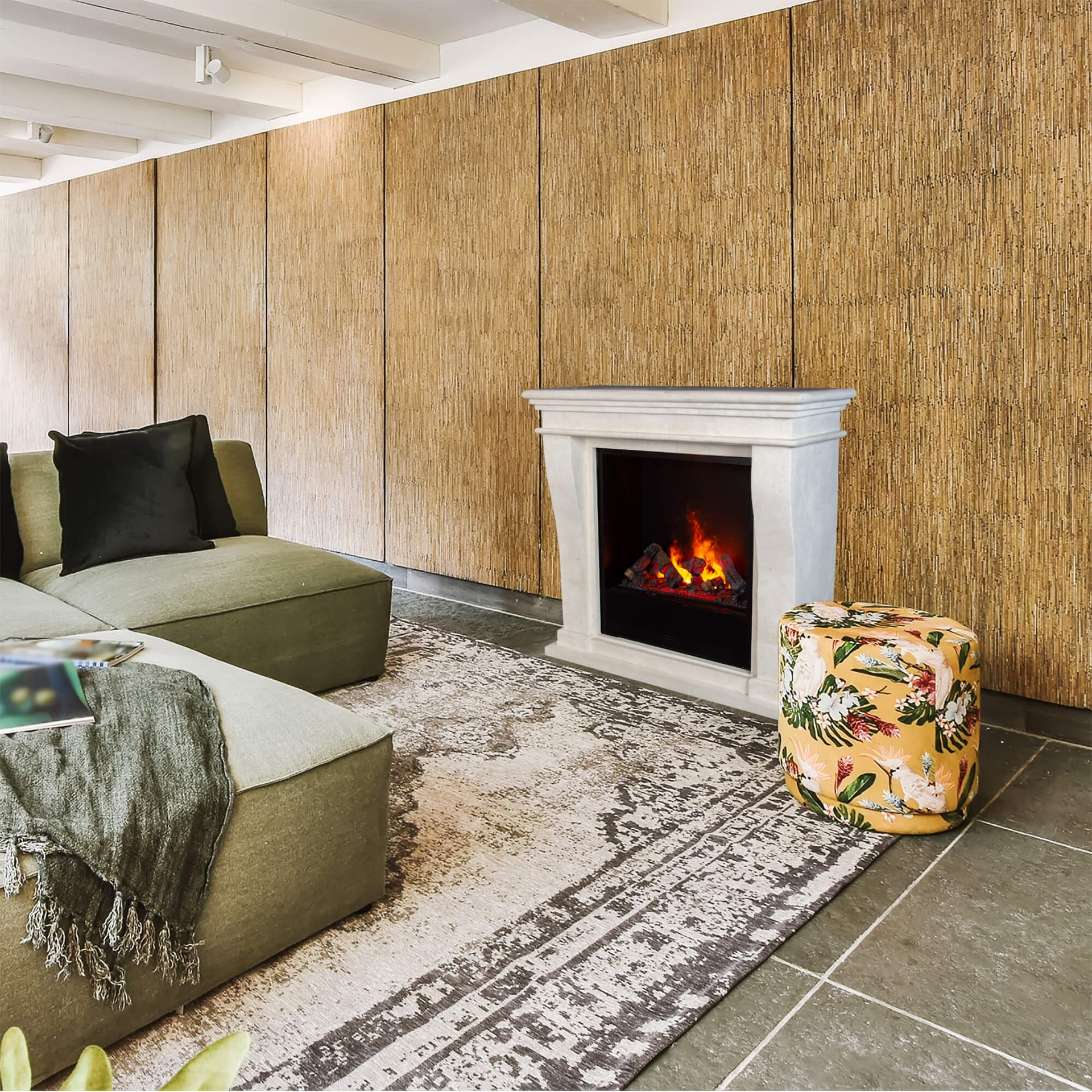 Een woonkamer met een groene bank, open haard, patroonkleed en bloemenhocker. De muur is van hout.