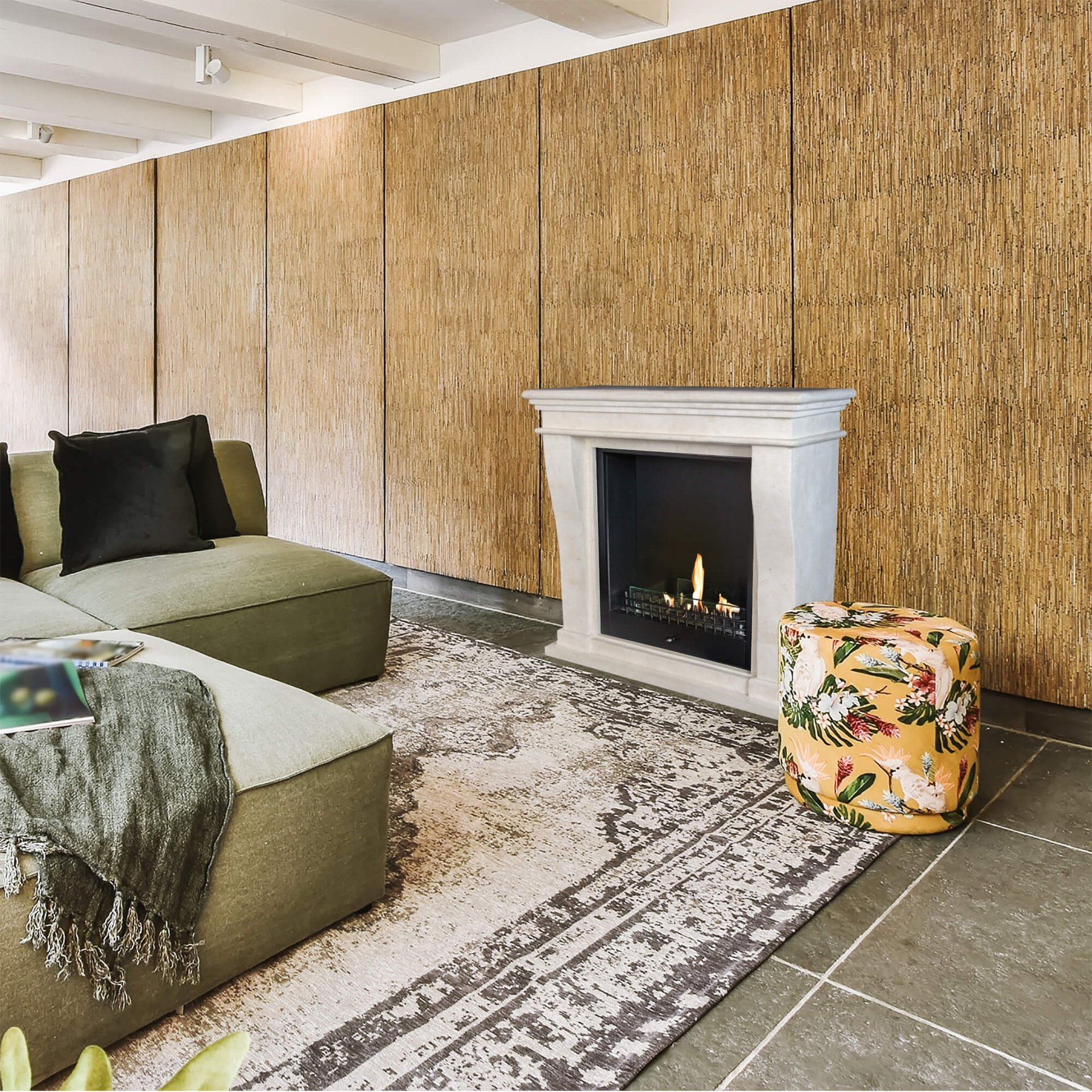 Een woonkamer met een olijfgroene bank, open haard, bloemenpoef en een patroonkleed. Houten panelen op de muur.