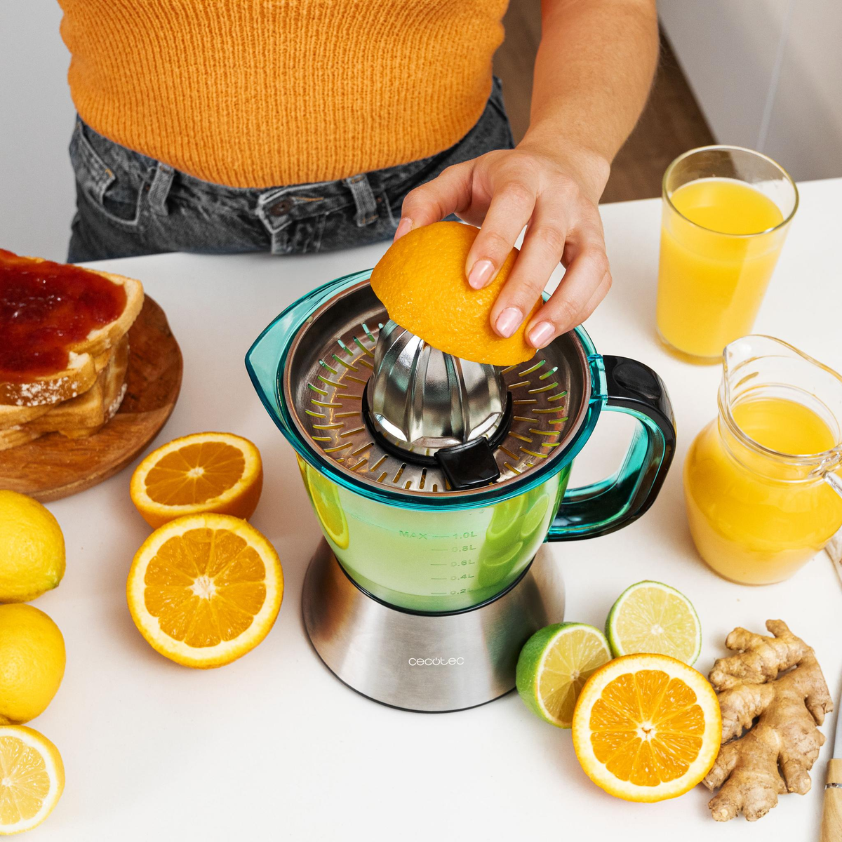 Persona exprimiendo naranjas con exprimidor, otras frutas y vaso de jugo en mesa blanca.