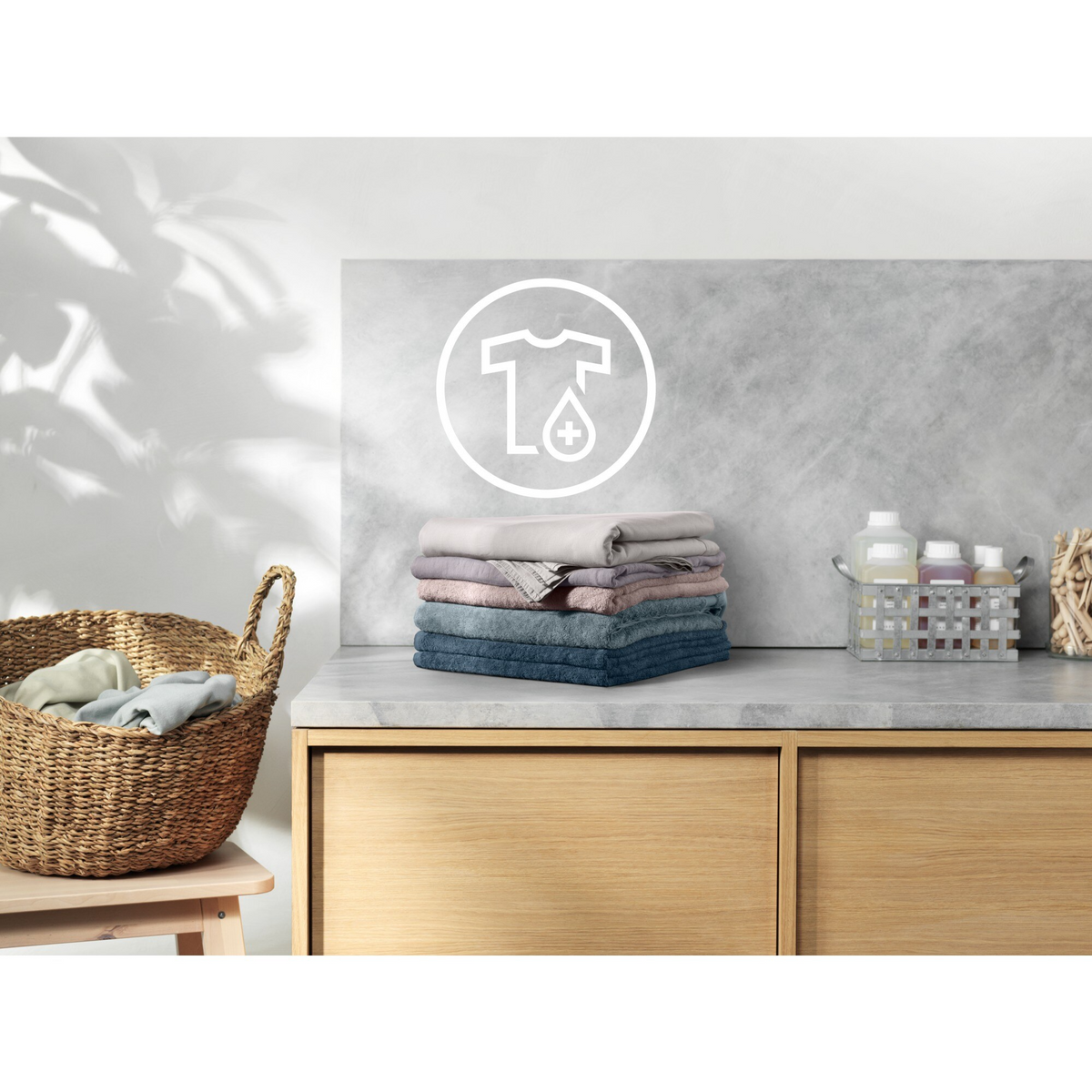 Stos złożonych ręczników na blacie, z symbolem prania powyżej. Kosz wiklinowy i butelki.