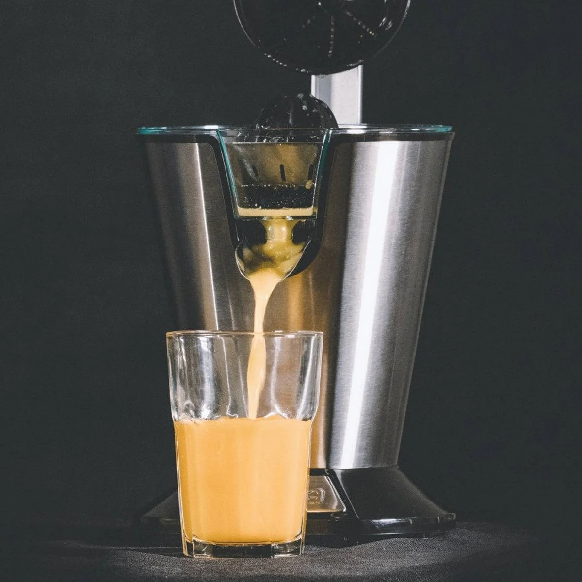 Spremiagrumi che versa succo d'arancia in un bicchiere. Design in acciaio inox e nero. Sfondo scuro.