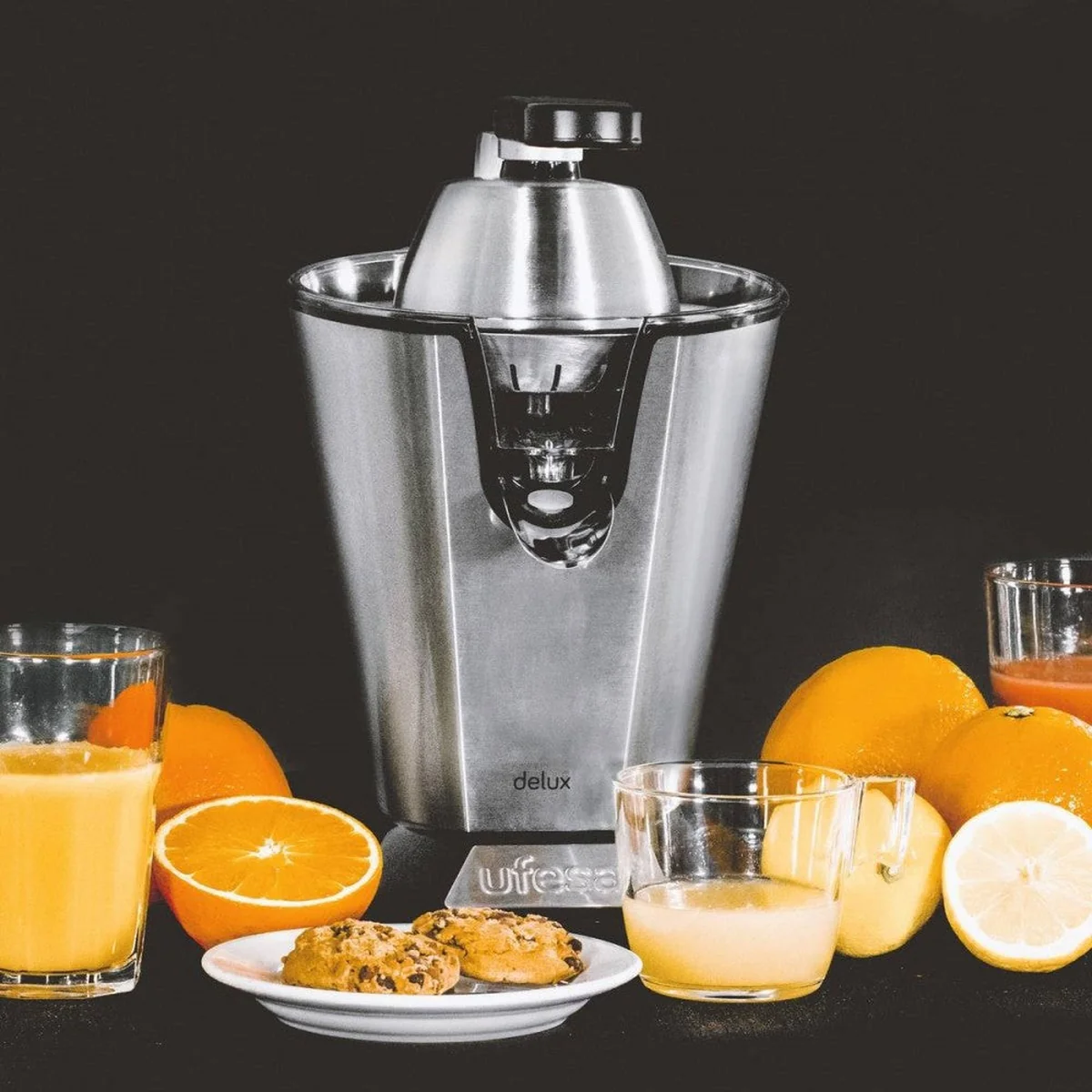 Centrifuga in acciaio inox con arance, bicchieri di succo e biscotti su una superficie nera.