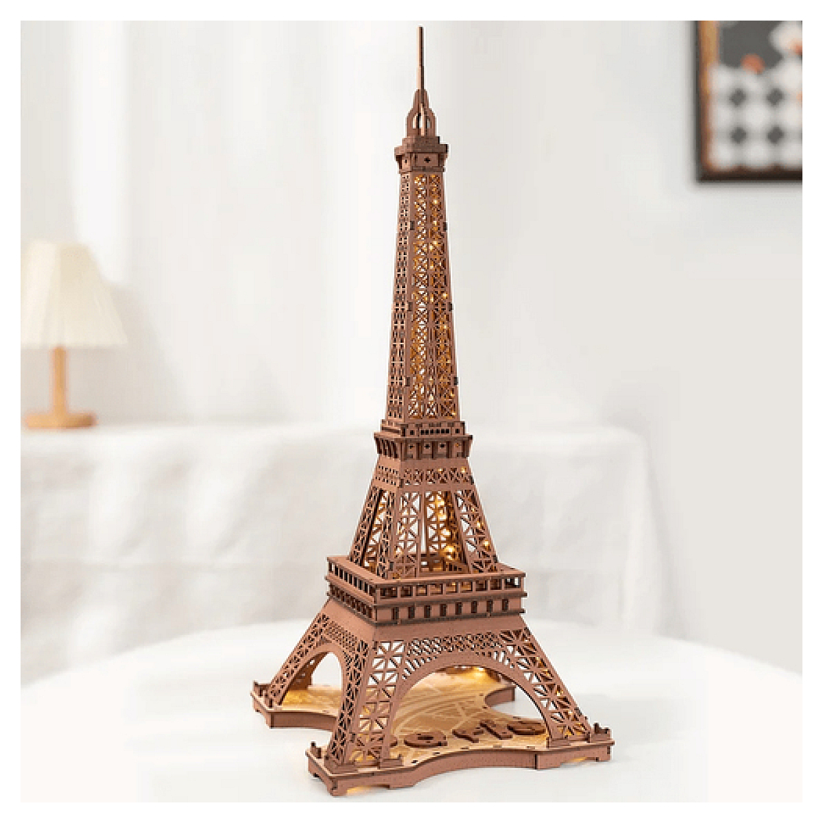 Podświetlana drewniana lampa w kształcie wieży Eiffla stoi na białej powierzchni, z jasną ścianą w tle.