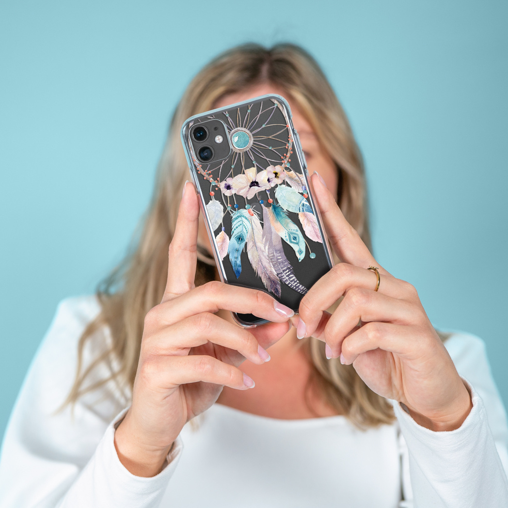 Femme tenant un téléphone avec une coque attrape-rêves, la personne porte une chemise blanche, fond bleu.