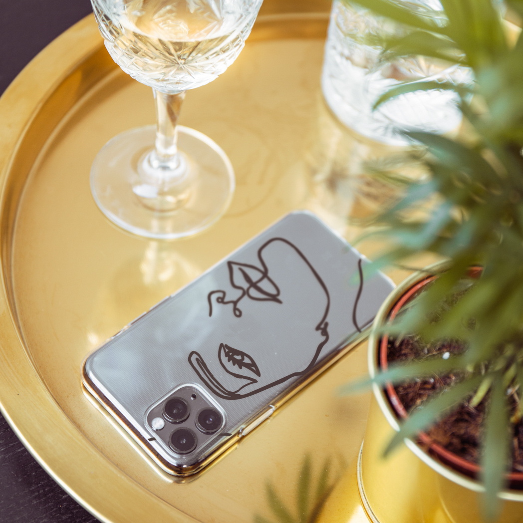 Téléphone avec étui en forme de visage sur un plateau doré, à côté d'un verre de vin et d'une plante en pot.
