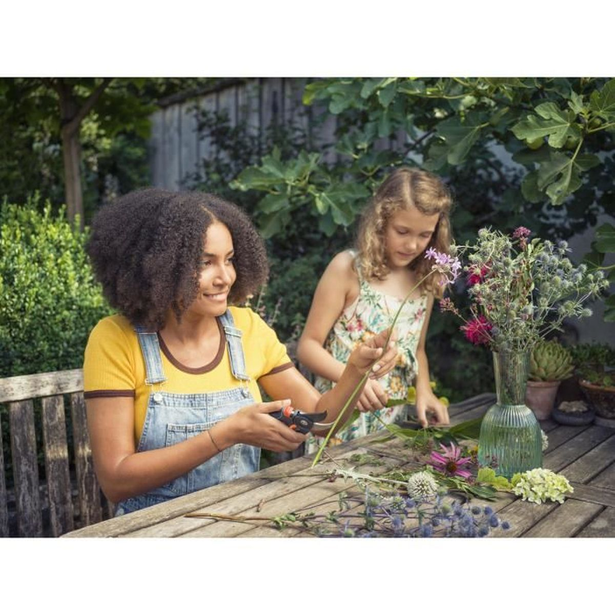 Dwie kobiety układają kwiaty. Stół z kwiatami i narzędziami. Zieleń w tle.