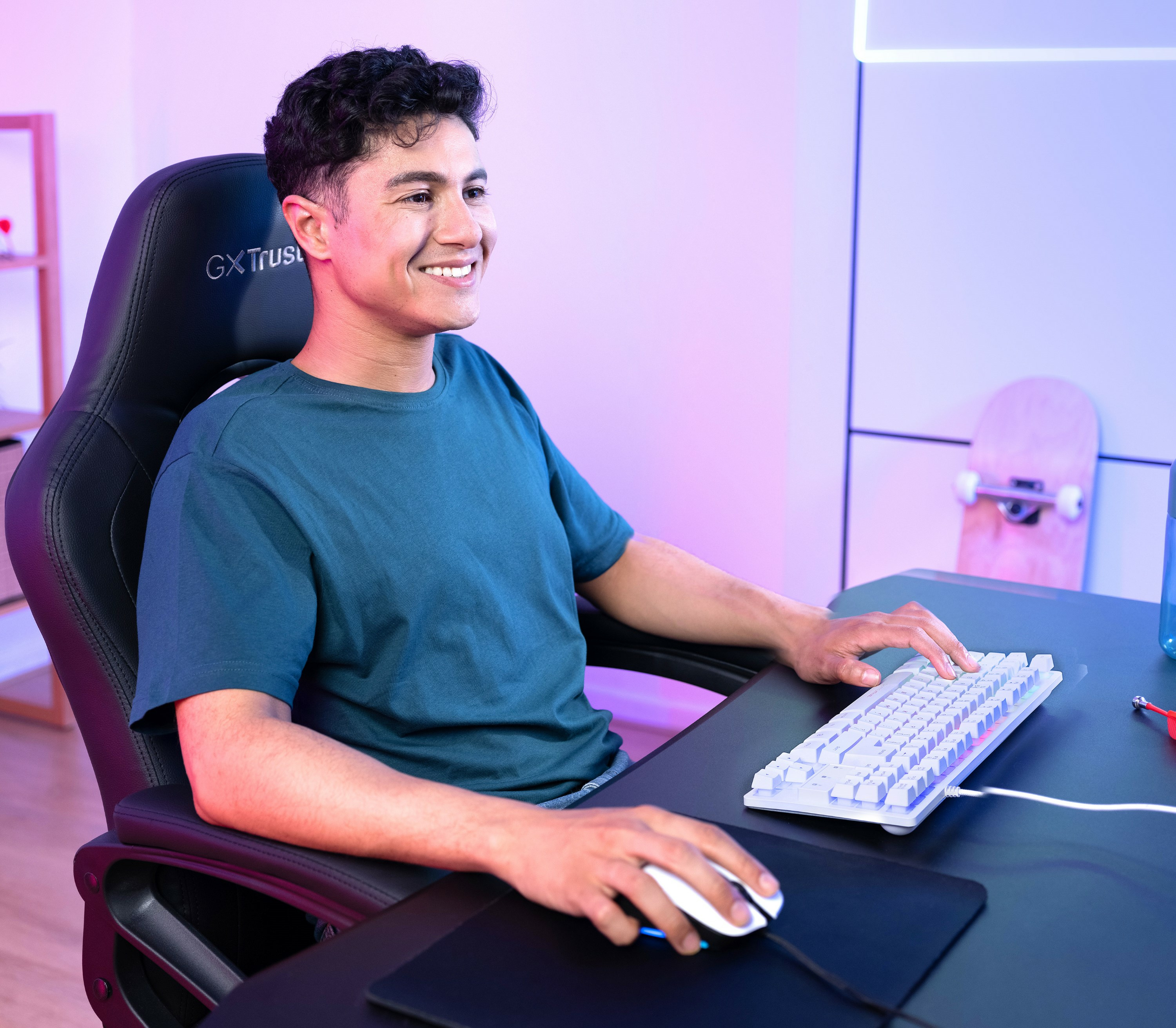 Uśmiechnięty mężczyzna przy biurku, używający białej klawiatury i myszy. Widoczny fotel do gier i deskorolka.