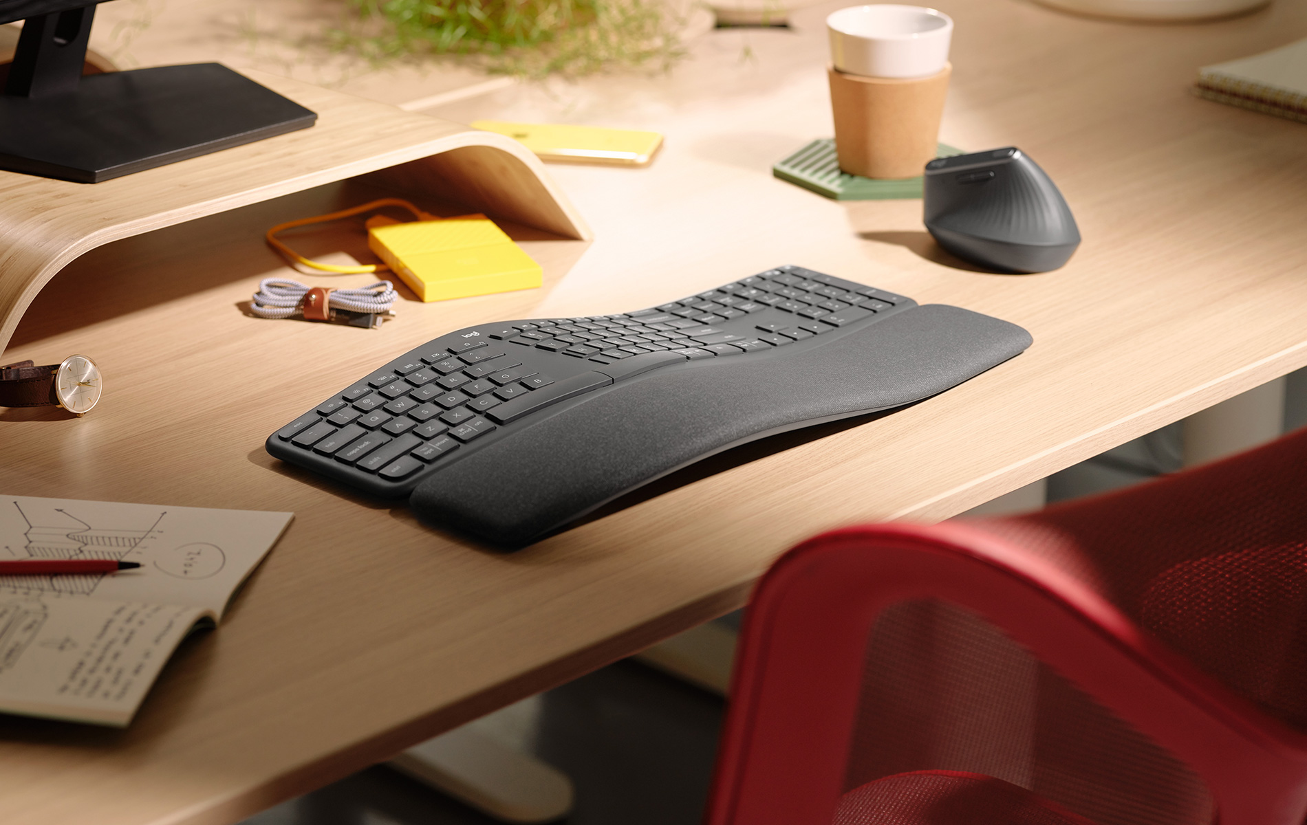 Tastiera ergonomica su una scrivania di legno, con un mouse, una tazza di caffè e una sedia rossa.