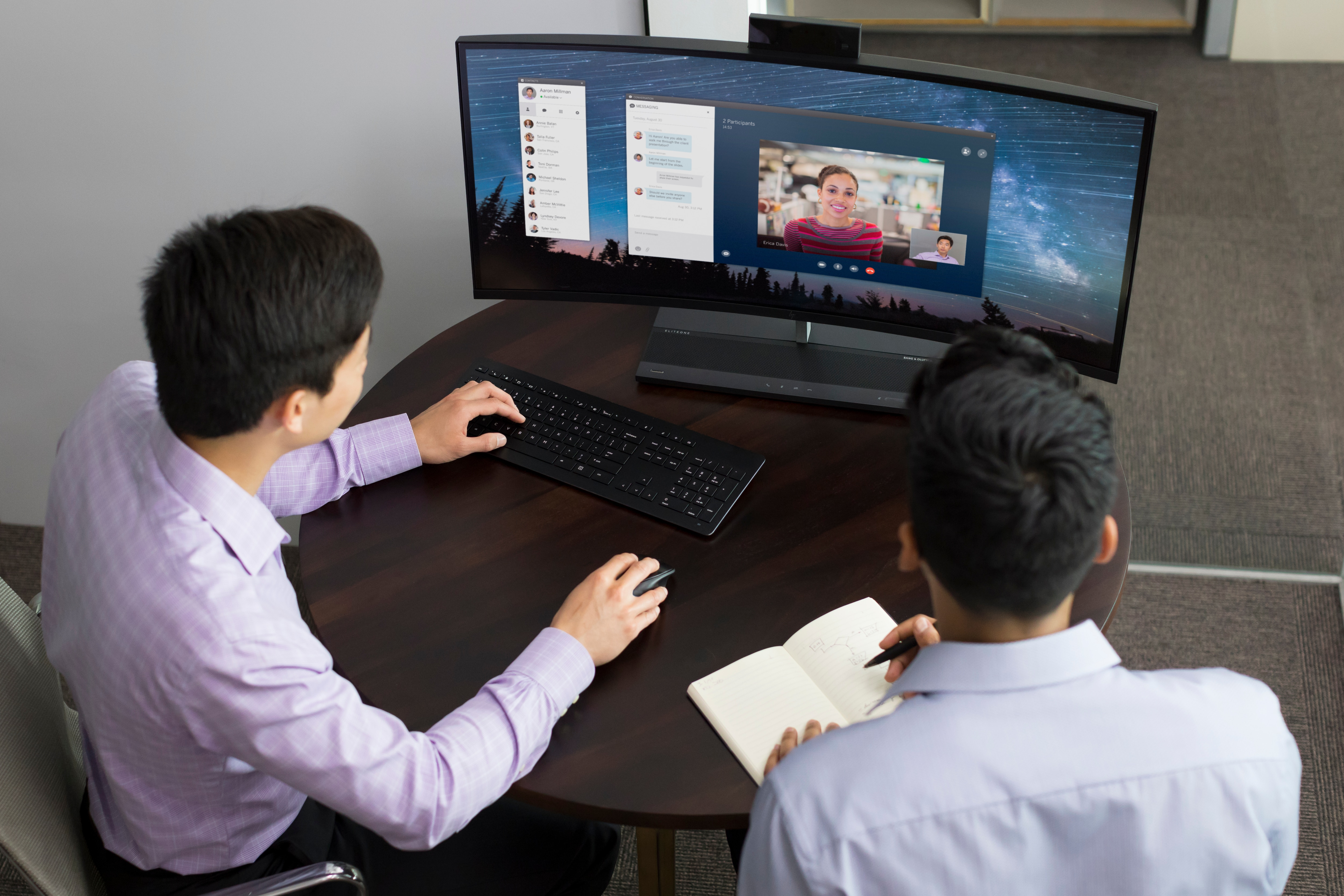 Dwóch mężczyzn przy stole, jeden używa klawiatury i myszy, drugi robi notatki na monitorze.