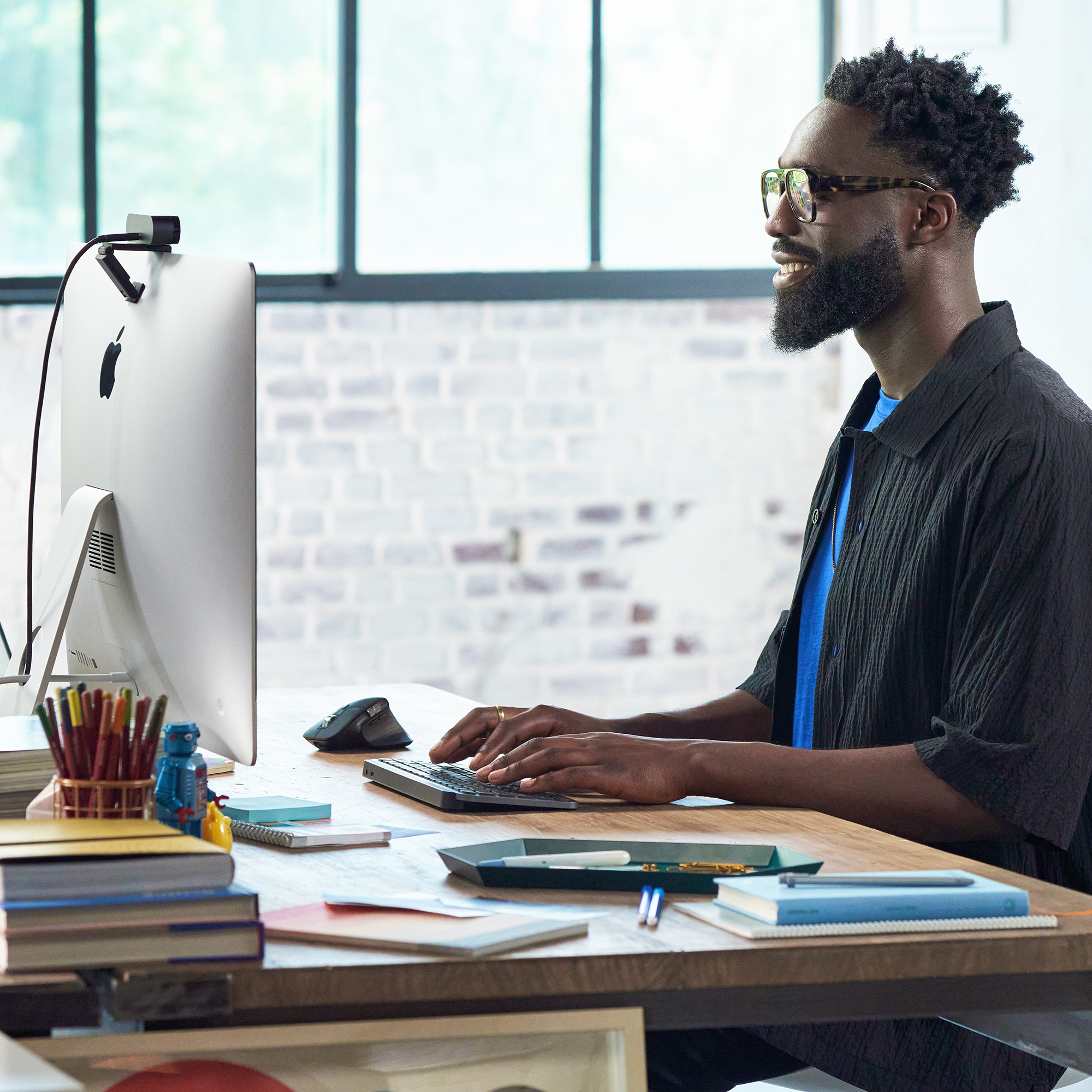 Mężczyzna w okularach pisze na klawiaturze obok iMaca. Są długopisy i książki. Tło to cegła.
