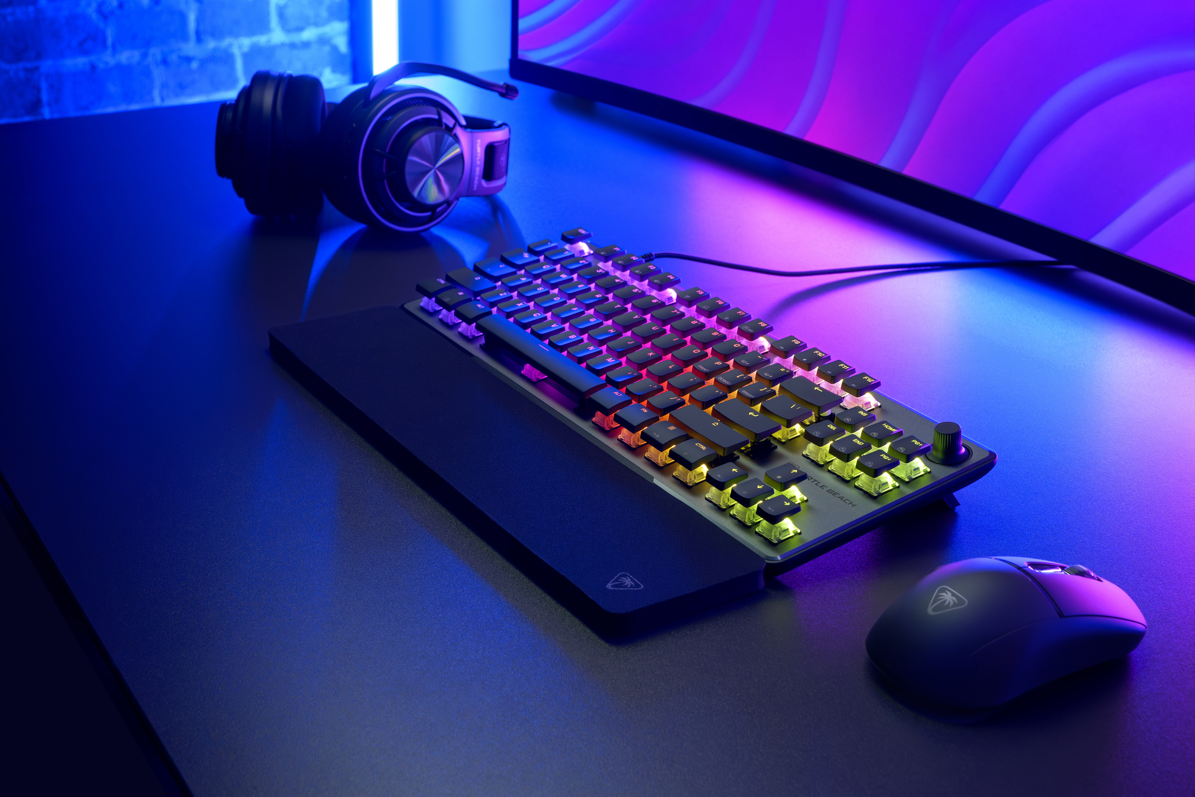Gaming-Setup mit Tastatur, Maus und Kopfhörern. Tastatur hat Regenbogenbeleuchtung. Monitor im Hintergrund.