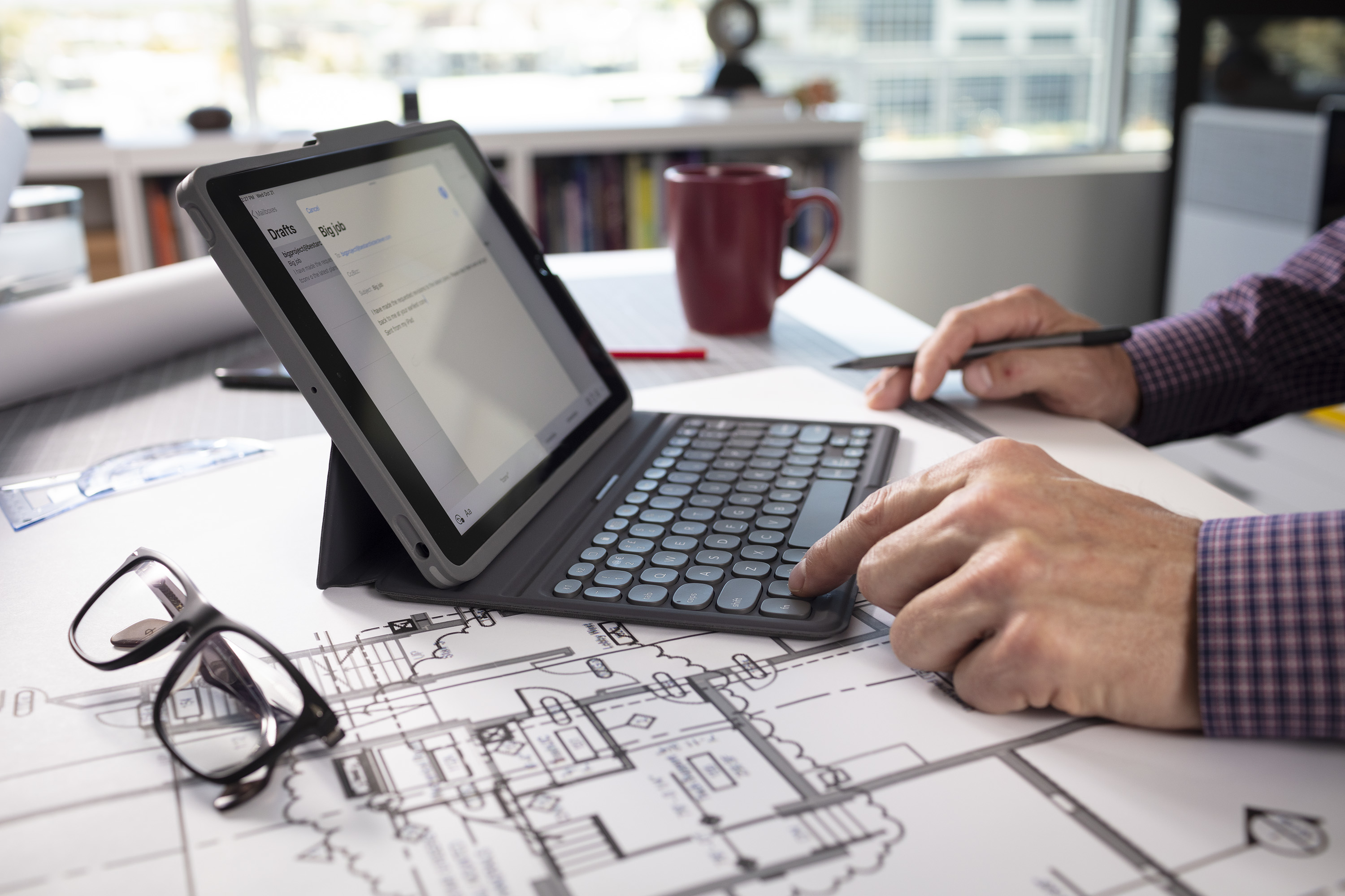 Eine Person benutzt ein Tablet mit Tastatur. Brille und eine Tasse sind im Vordergrund. Blaupausen sind sichtbar.