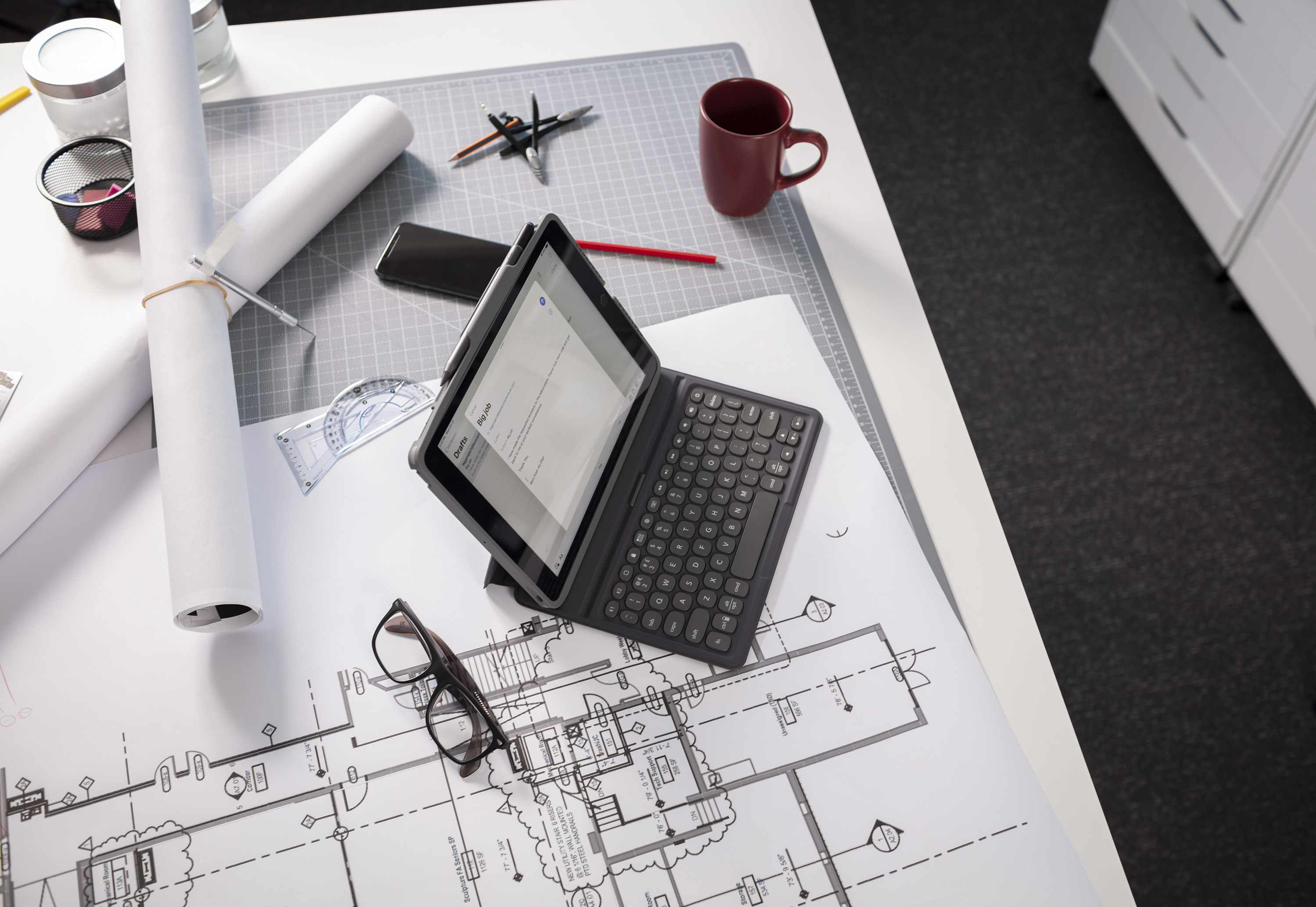 Tablet auf einem Bauplan, mit Brille, Bleistiften und anderen Arbeitswerkzeugen. Das Tablet ist geöffnet.
