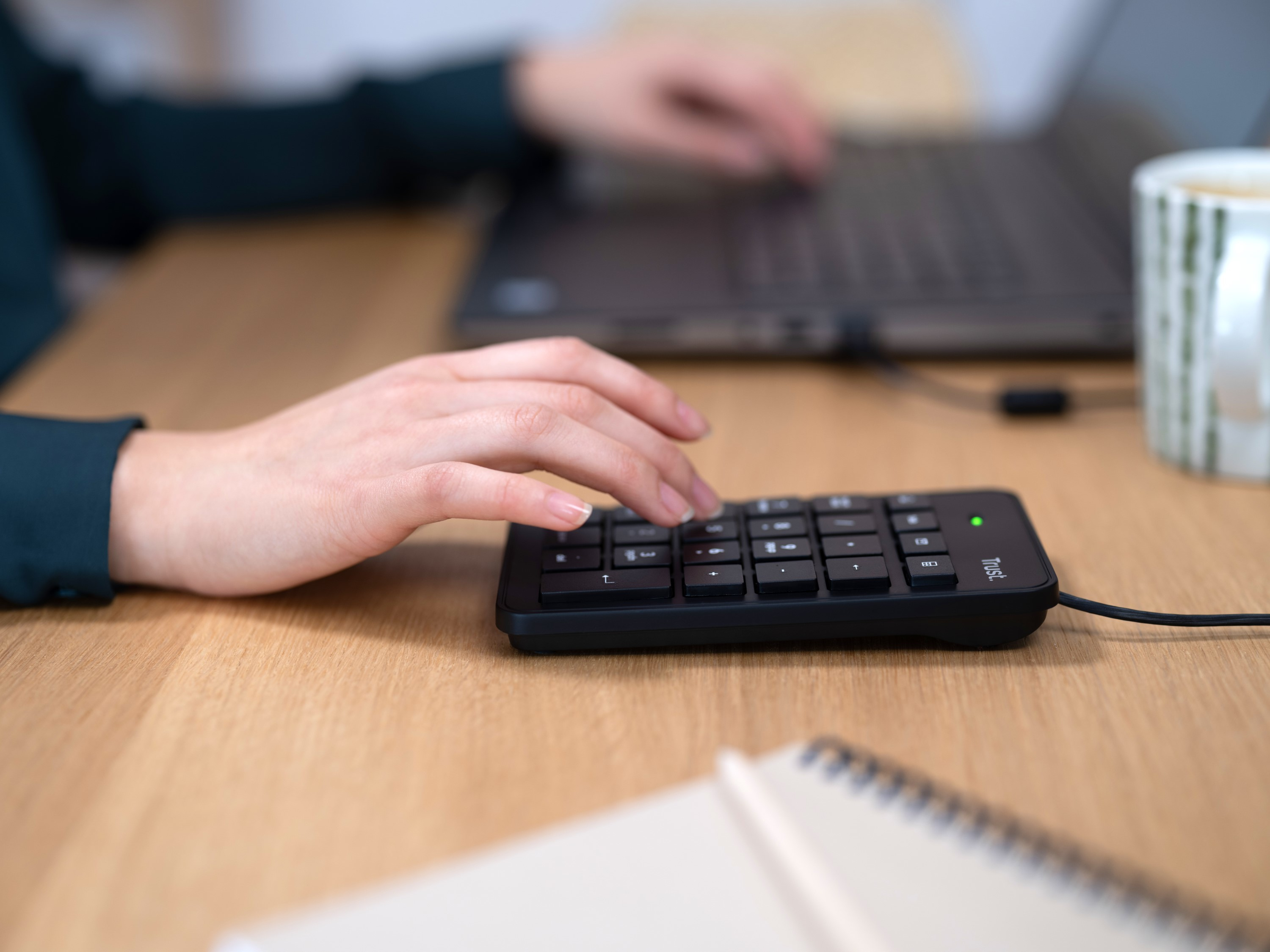 Die Hand einer Person, die einen schwarzen Nummernblock auf einem Holzschreibtisch neben einem Laptop und einer Tasse Kaffee benutzt.