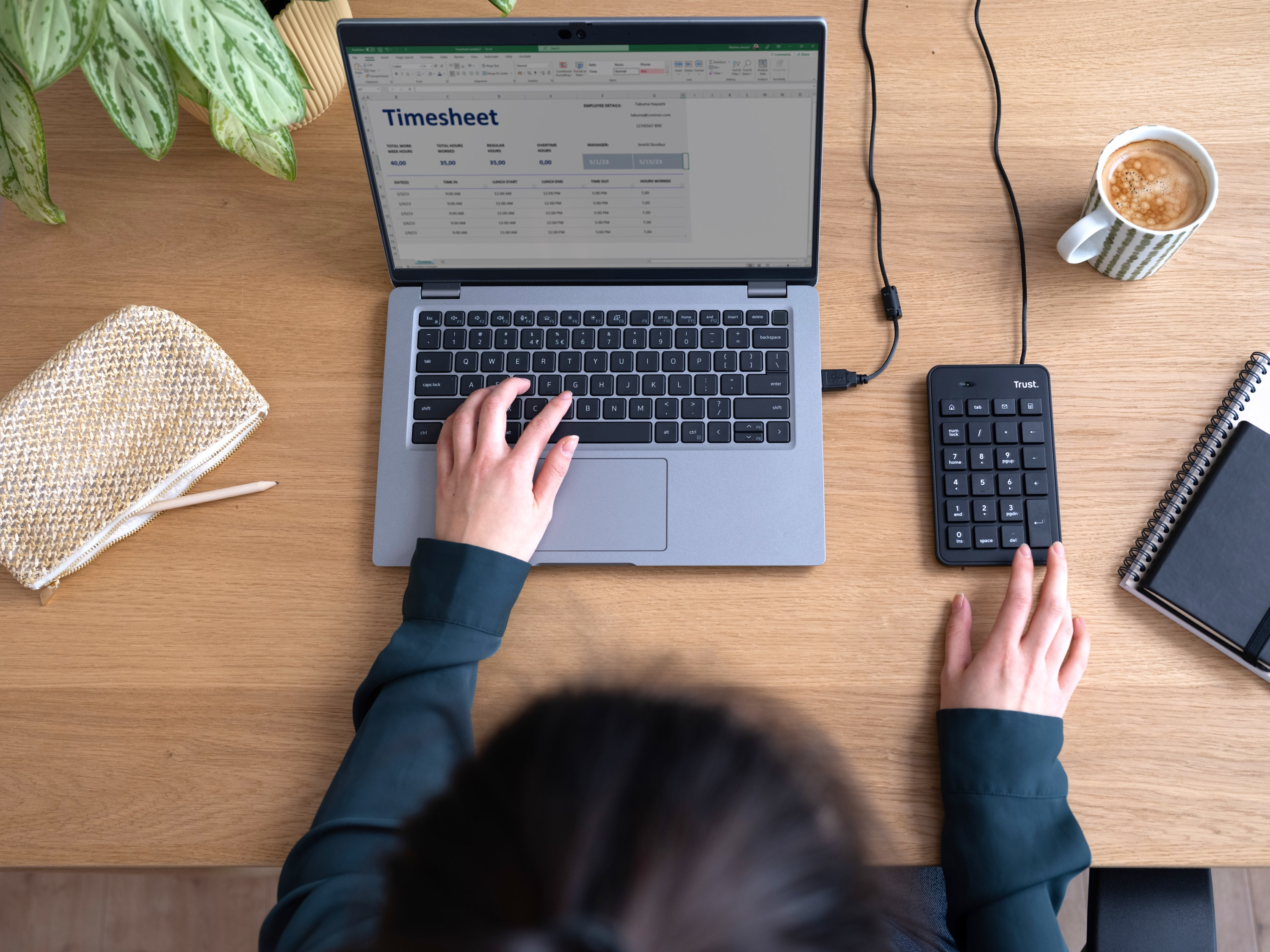 Eine Person arbeitet an einem Laptop mit einem angezeigten Arbeitszeitnachweis. Ein Nummernblock ist angeschlossen. Eine Tasse Kaffee ist vorhanden.